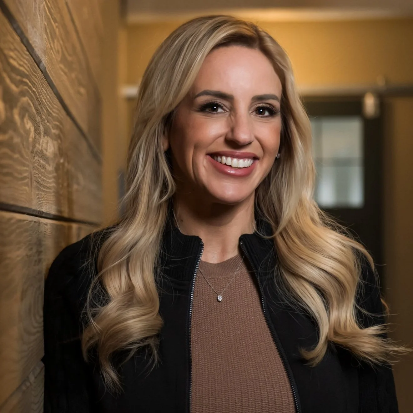 Smiling person with long blonde hair, wearing a dark jacket and brown top, standing indoors against a wooden wall.