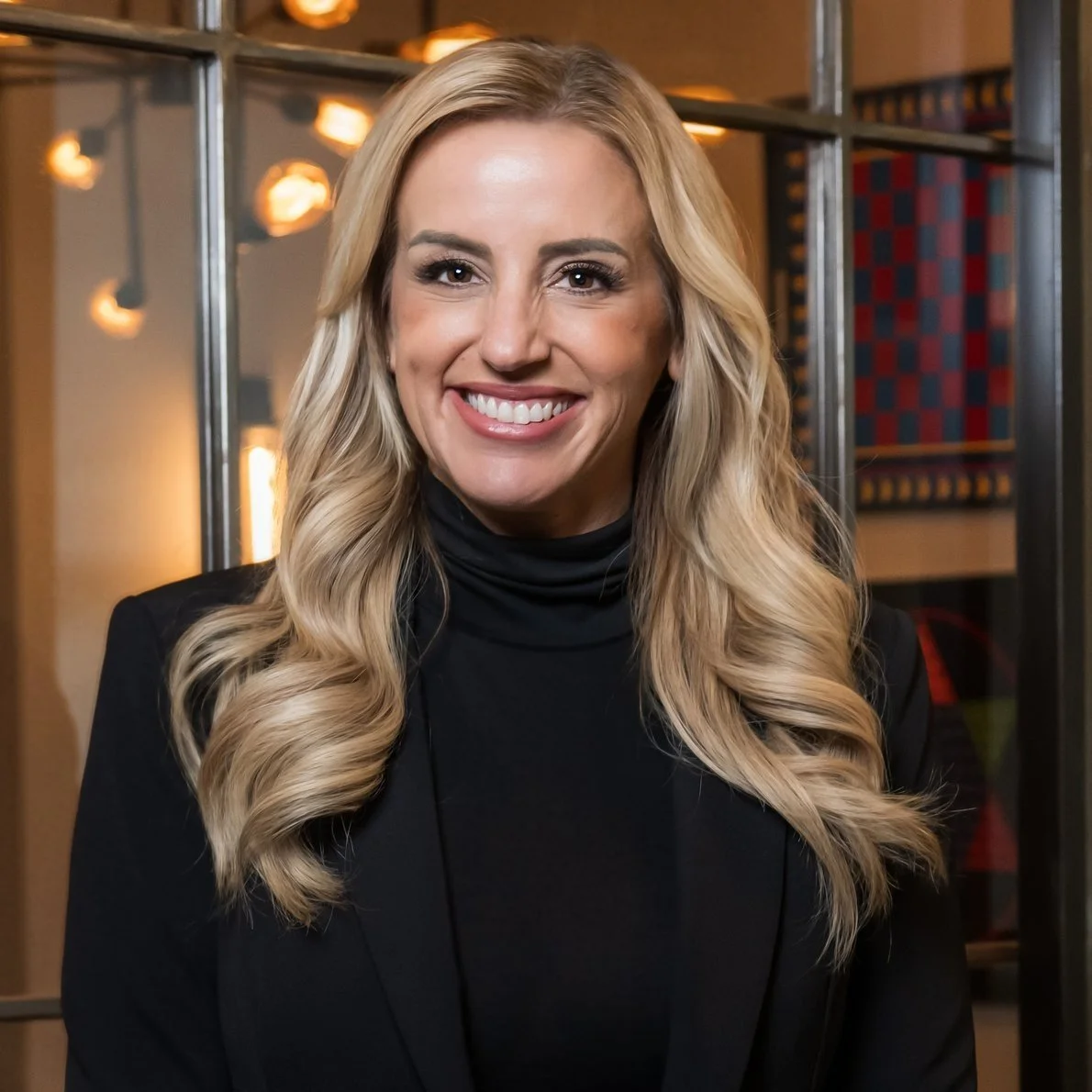 A woman with long blonde hair wearing a black turtleneck and blazer, smiling in front of a background featuring lights and a window reflection.