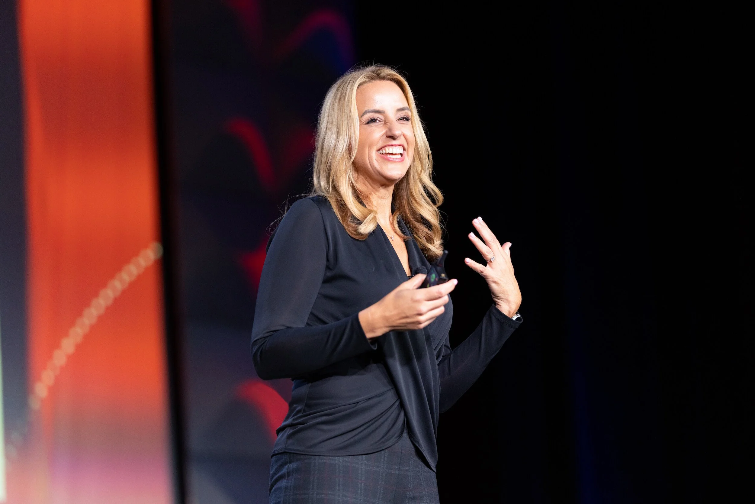 Person speaking on stage with a microphone, wearing a black blouse, against a dark background with orange accents.