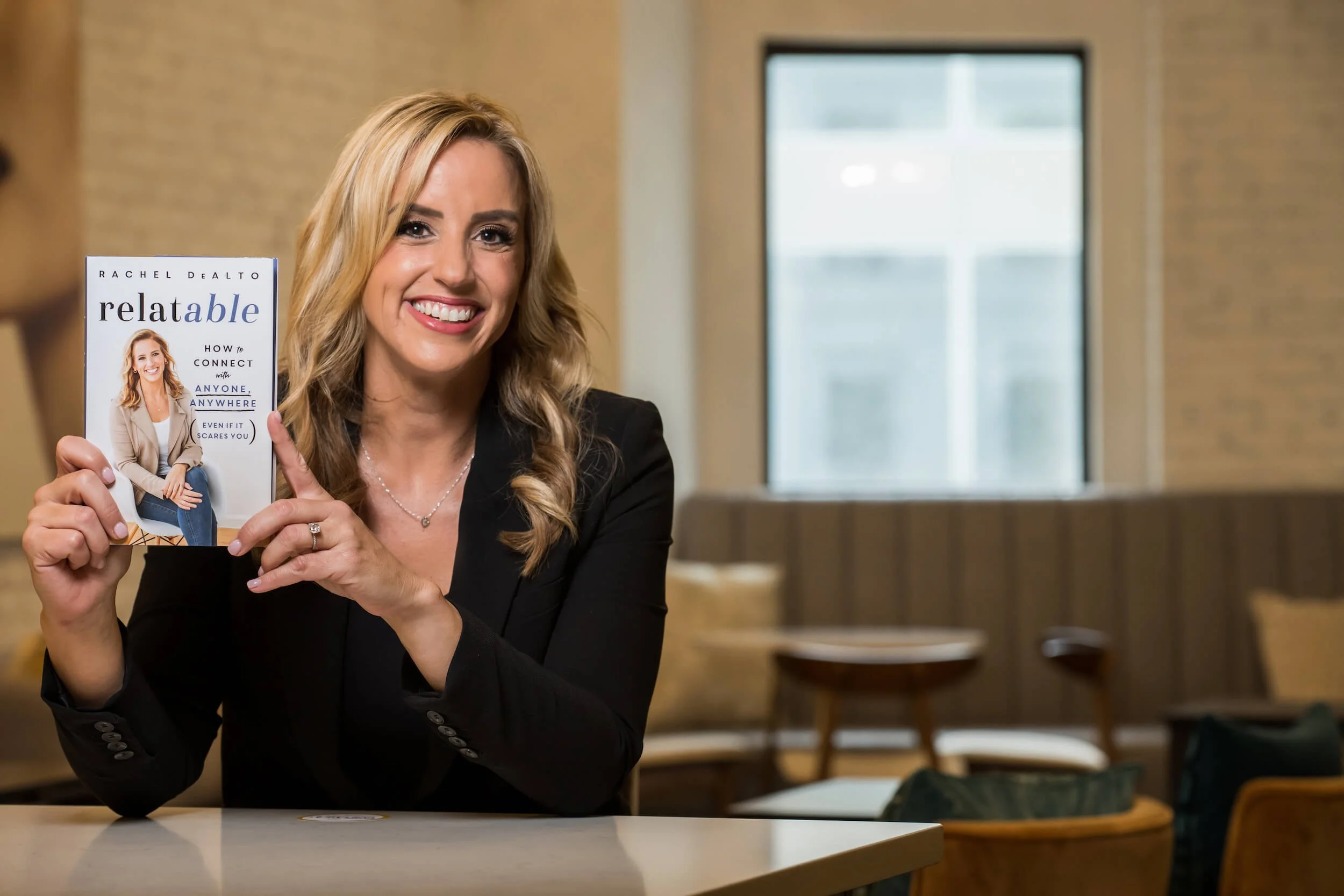 Person in a black blazer holding a book titled 'relatable' with a smiling face on the cover, in a cozy indoor setting with modern furniture.
