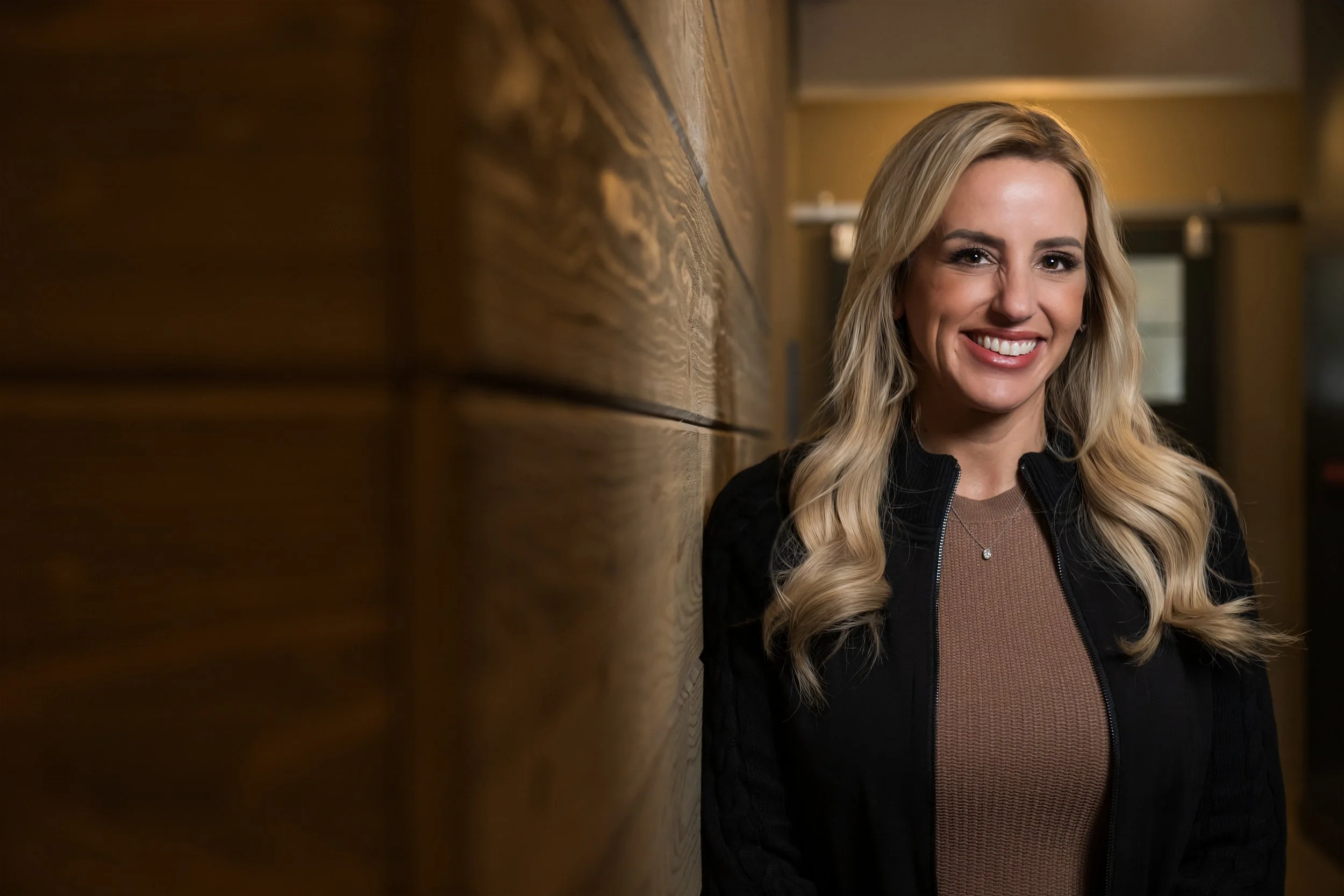 Smiling woman with long, wavy blonde hair, wearing a black jacket and a brown top, leaning against a wooden wall in an indoor setting.