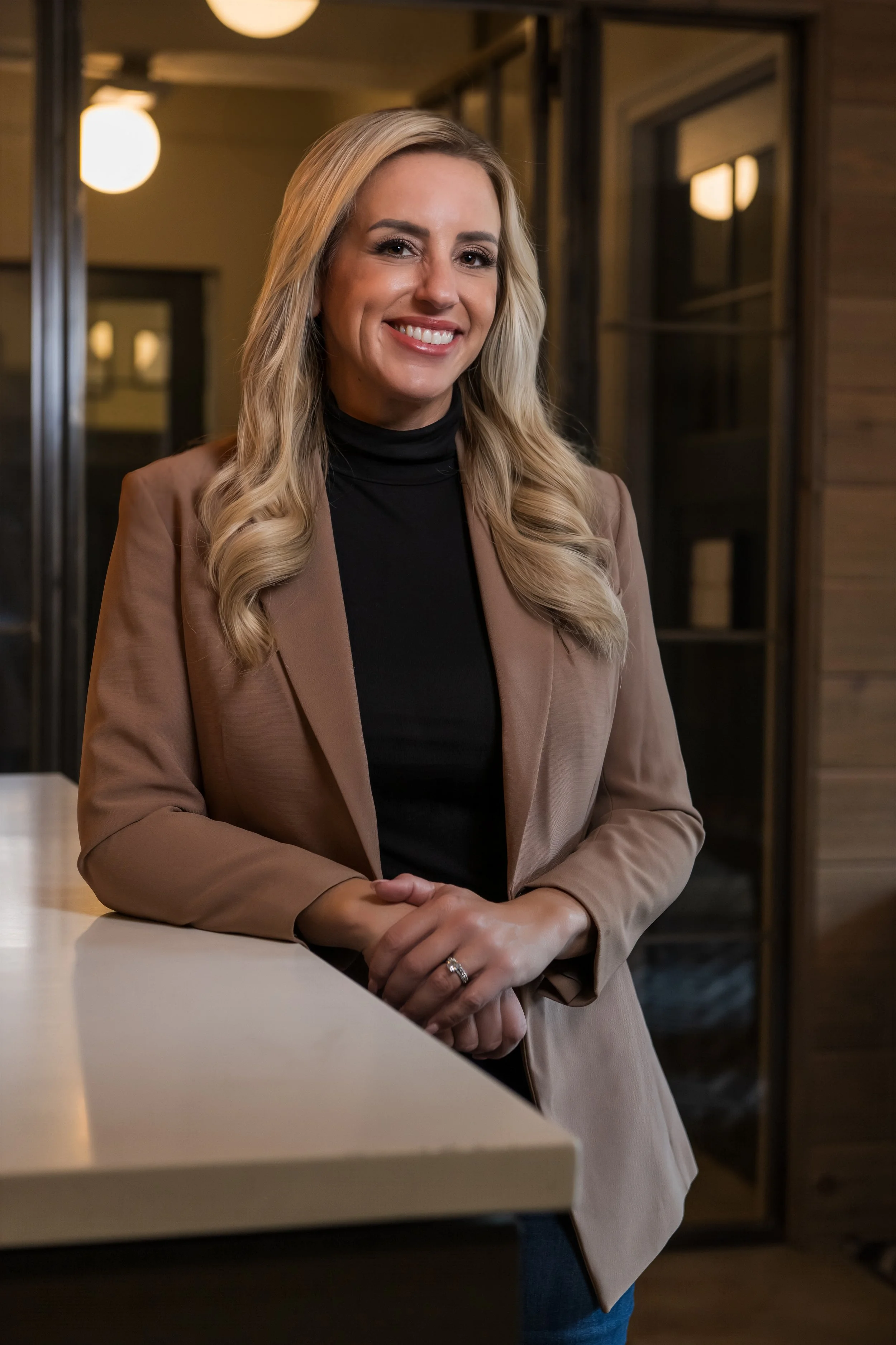 A woman with long blonde hair, wearing a black turtleneck and beige blazer, smiling and leaning on a beige countertop in an indoor setting with warm lighting and wooden visible walls.