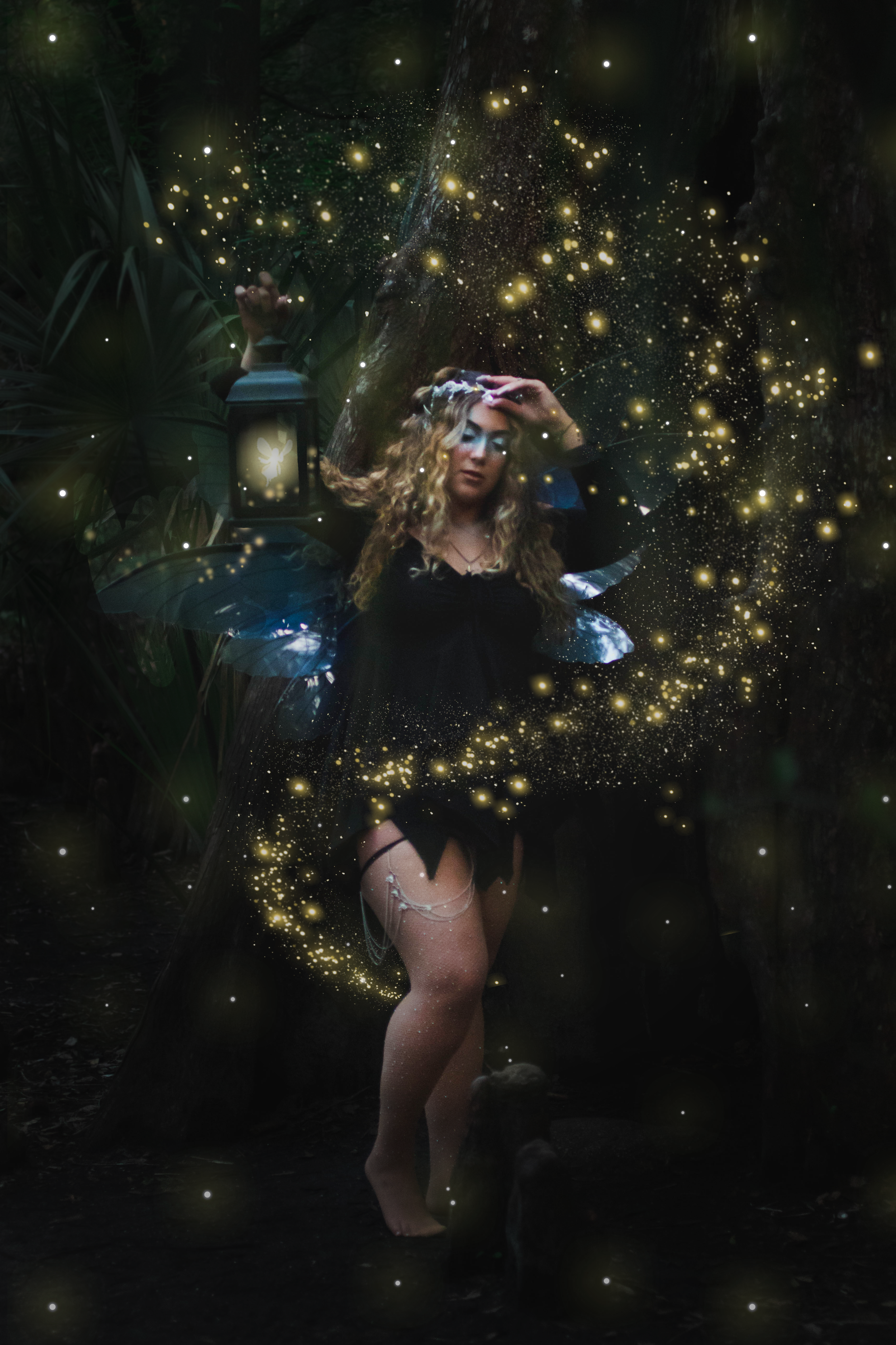 A woman dressed as a fairy with fairy wings, surrounded by glowing fireflies, standing in a dark forest setting. She is positioned next to a lantern with a bunny silhouette, and she is touching her forehead while looking down, creating a magical and 