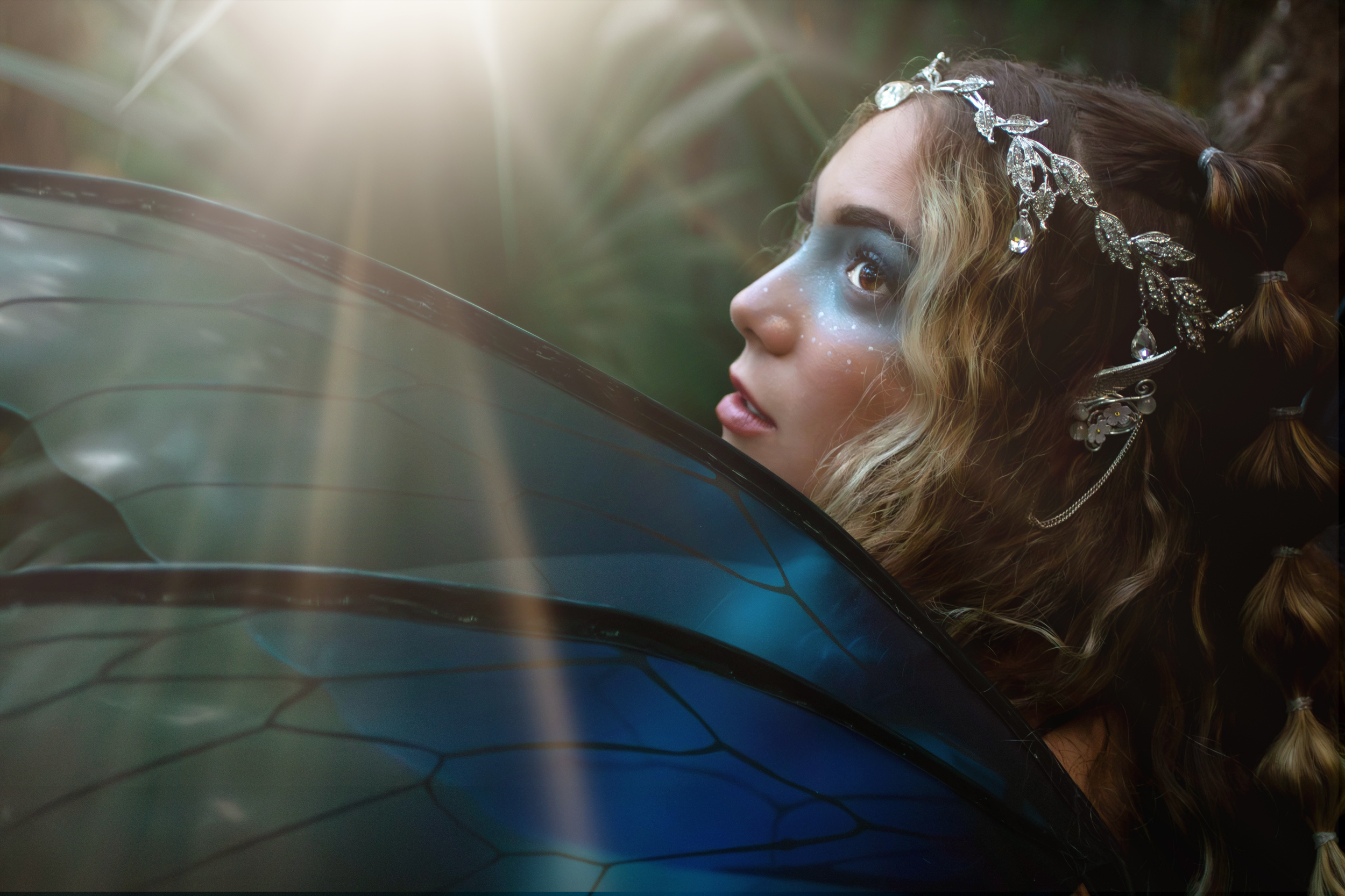 A woman with makeup and a decorative silver headpiece looking to the side, surrounded by green foliage with sunlight filtering through.