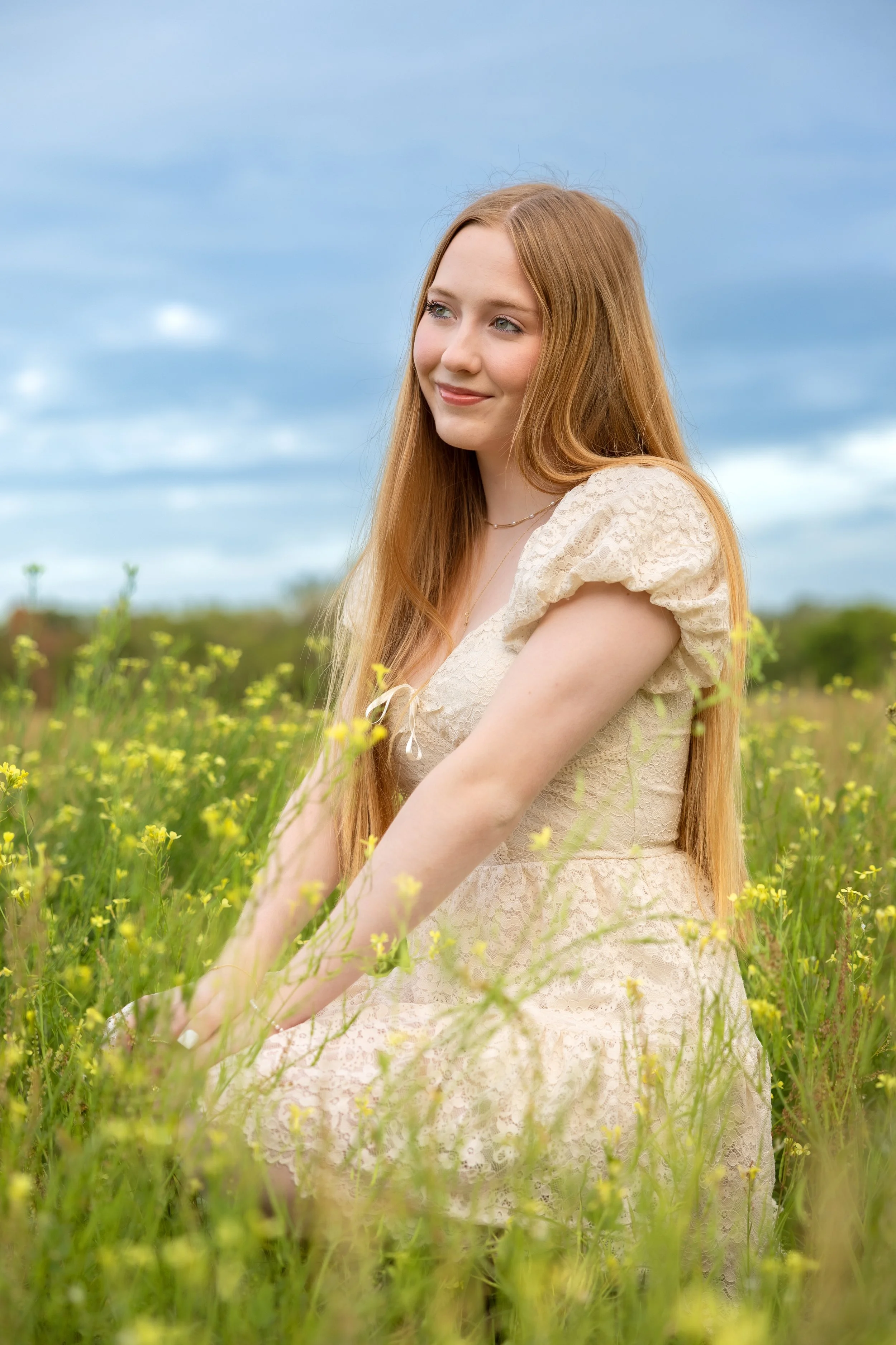 A young woman with long red hair wearing a cream dress standing in a field of yellow flowers under a cloudy sky.