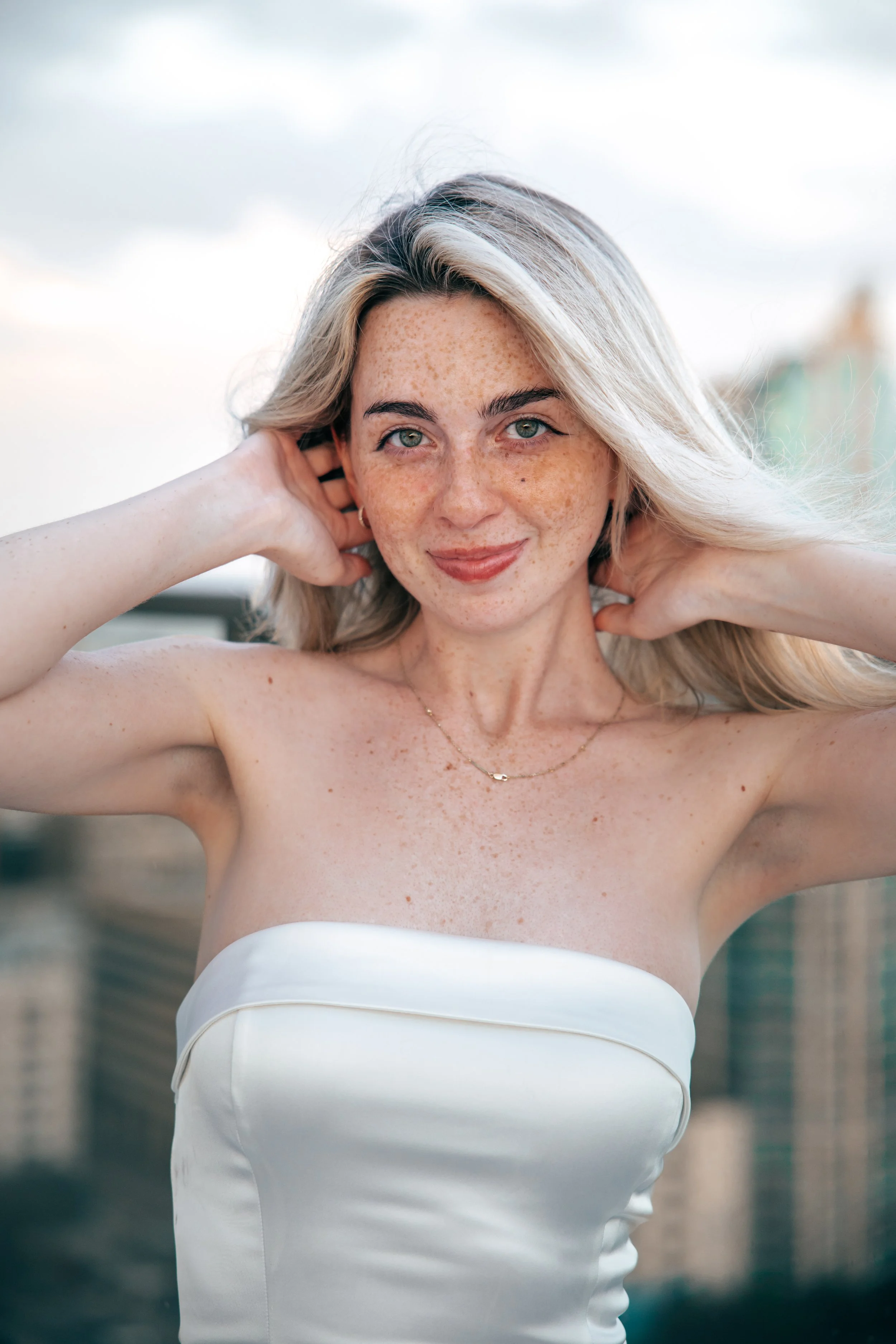 A woman with light blonde hair, blue eyes, and freckled skin posing outdoors, wearing a strapless white dress and a delicate gold necklace, with her hands behind her head and a soft smile.