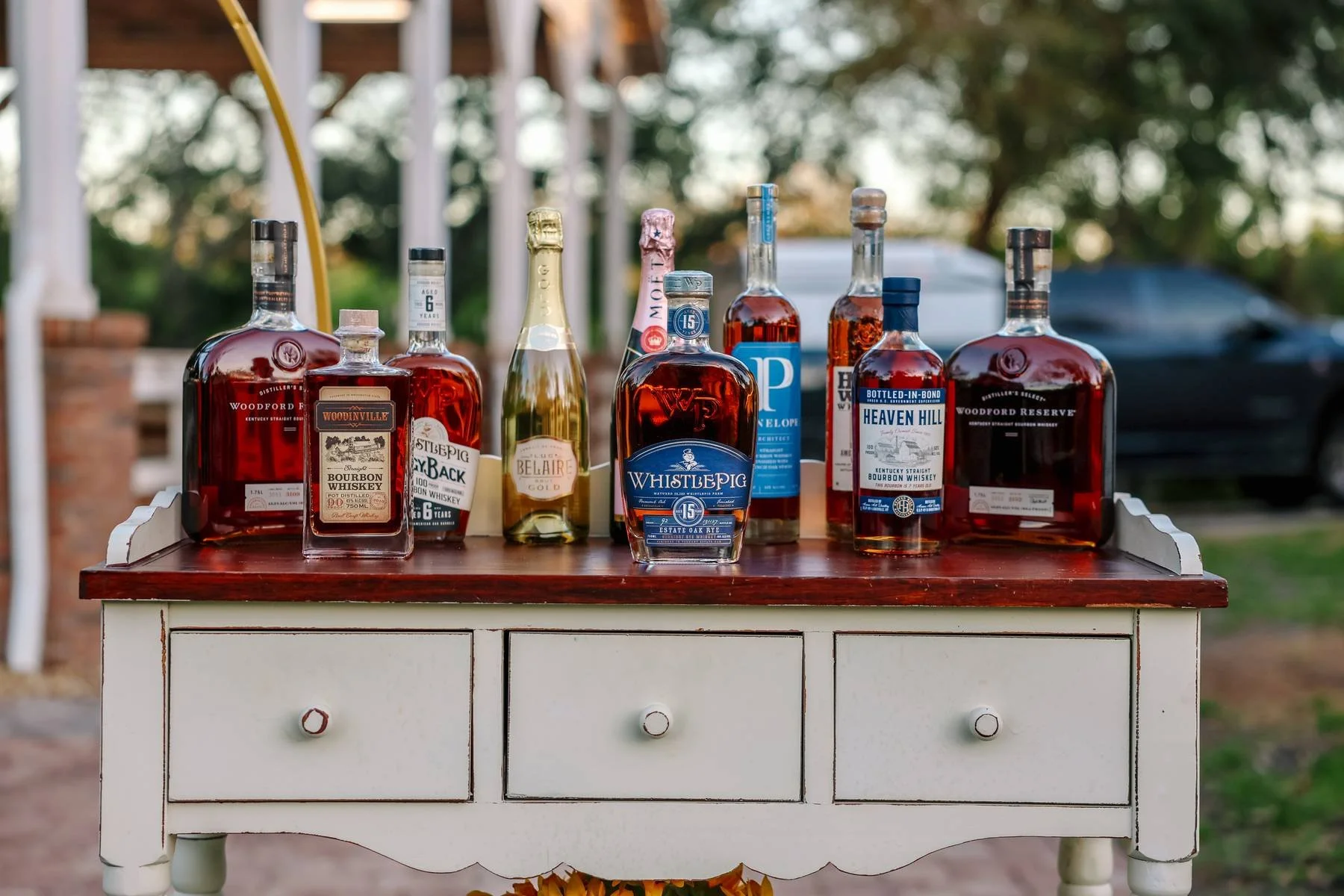 Assorted bottles of whiskey and bourbon displayed on a white wooden table with drawers, outdoors with blurred trees in the background.