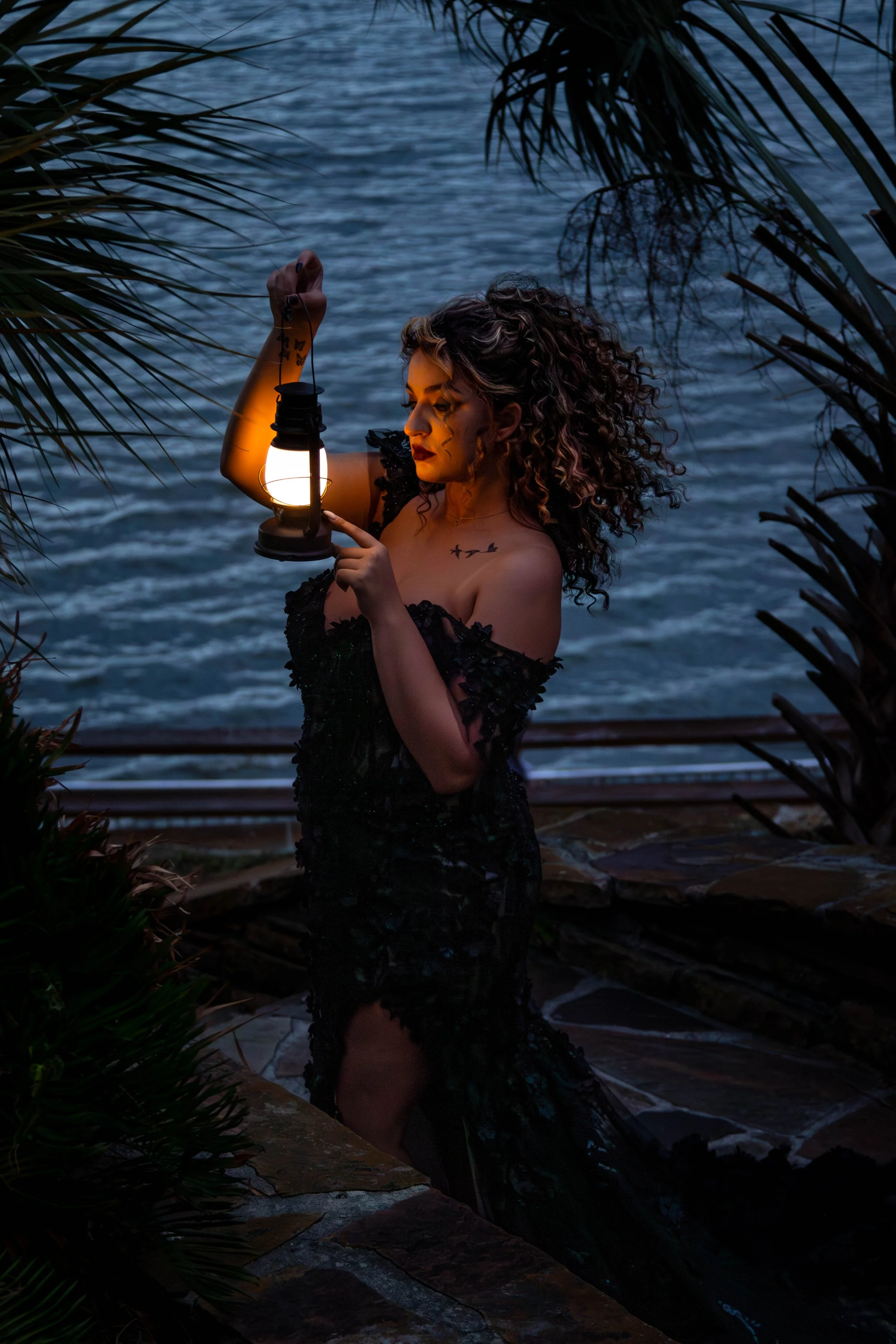 A woman with curly hair, wearing a black lace dress, holding a lantern, standing by water surrounded by leafy plants in low lighting.