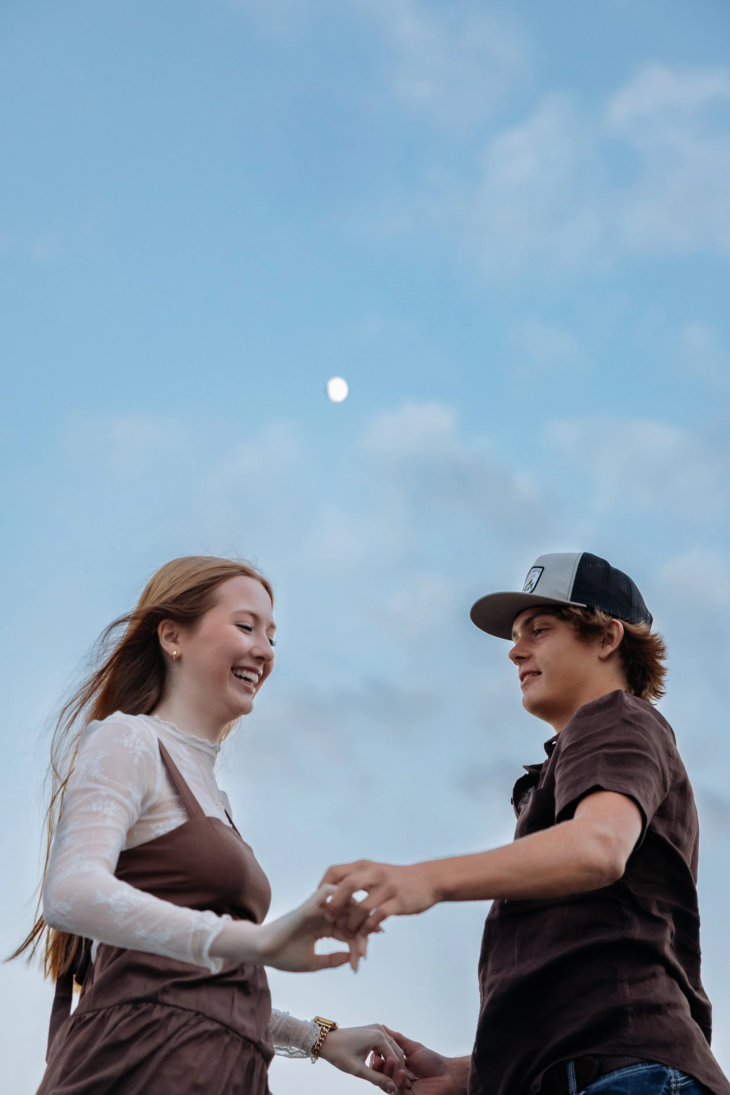 A young couple holding hands and smiling outside under a blue sky with clouds and a visible moon.
