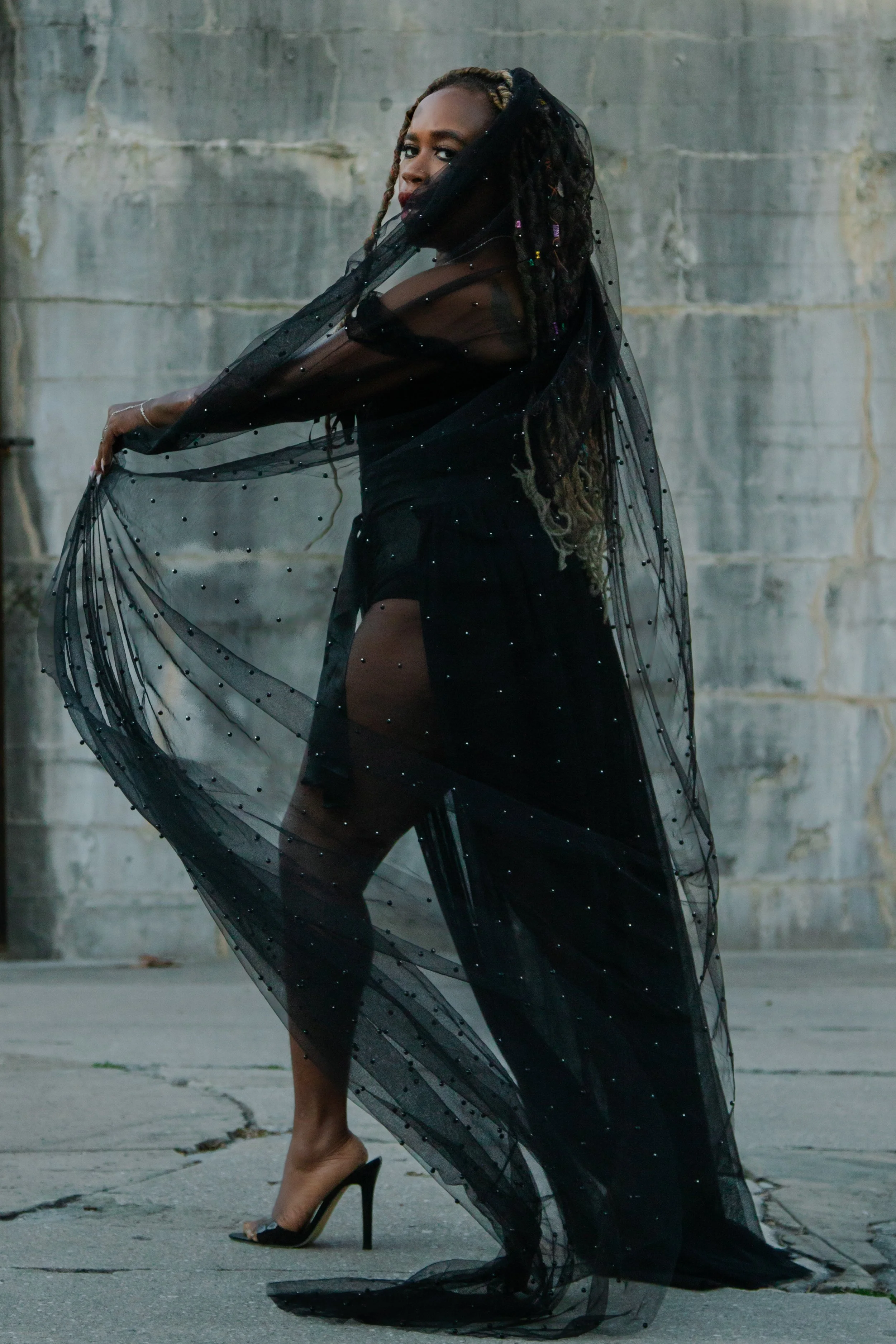 A woman with long, braided hair wearing a sheer black dress with colorful embellishments, holding a sheer black cape, standing outdoors in front of a concrete wall.