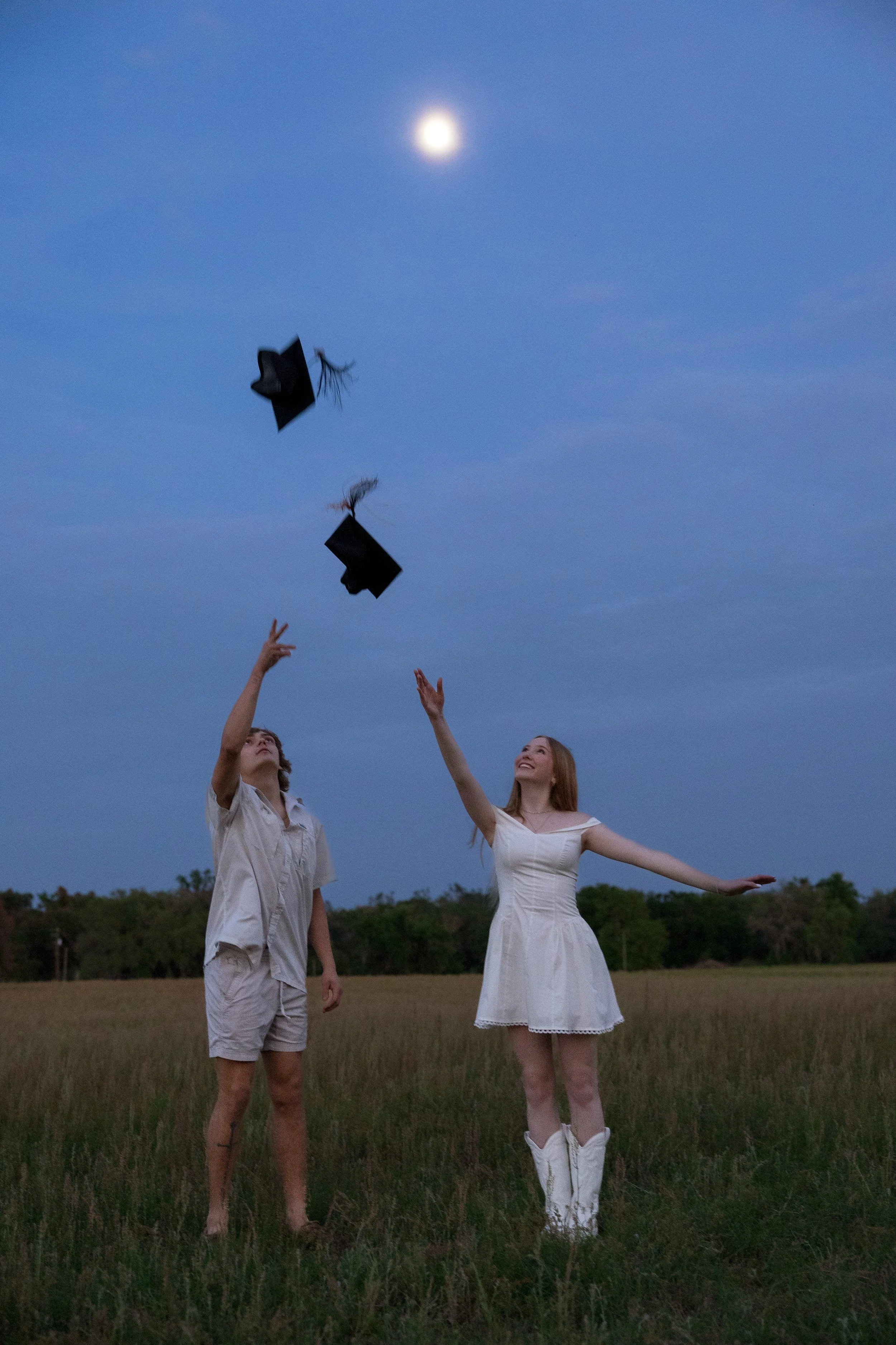 Two young women in white dresses and boots standing in a field at dusk, tossing graduation caps into the air with the moon in the sky above.