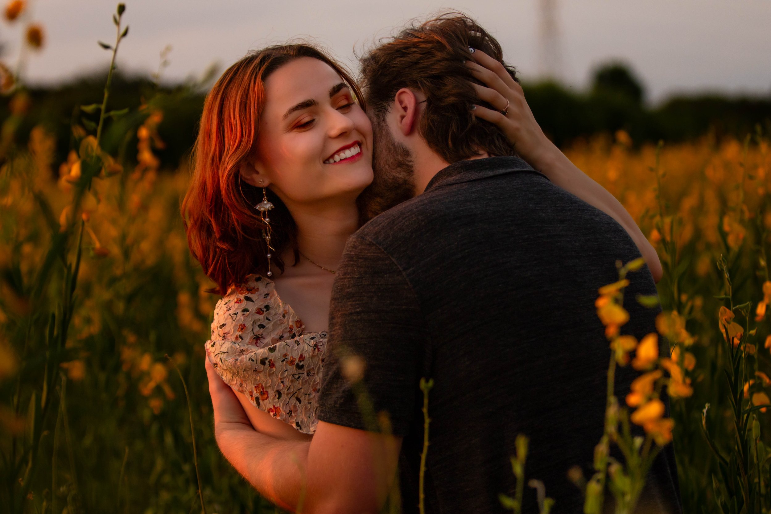 A couple embracing in a field of yellow flowers during sunset. The woman has red hair, is smiling, and is wearing floral earrings and a floral top. The man has brown hair and a beard, with his face turned away.