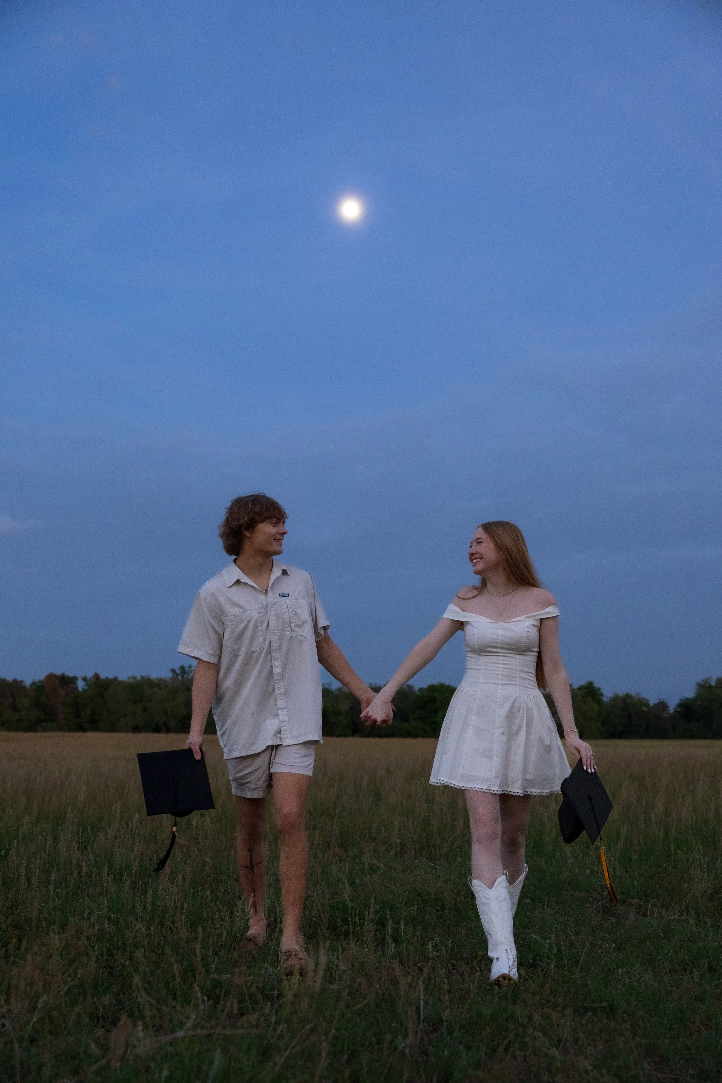 A young man and woman holding hands and walking through a field during twilight, with a full moon visible in the sky and tree line in the background.