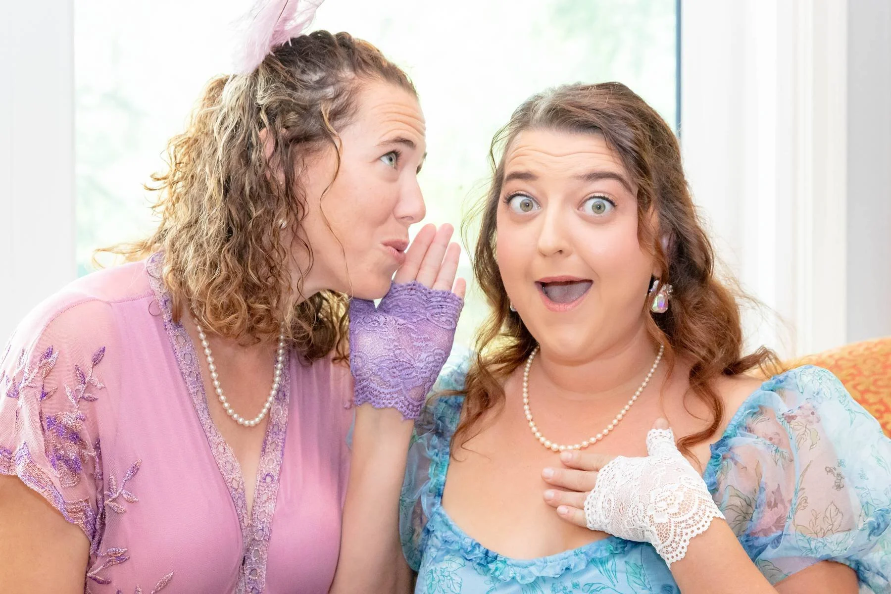 Two women in colorful costumes, one whispering to the other who appears surprised, inside a bright room.