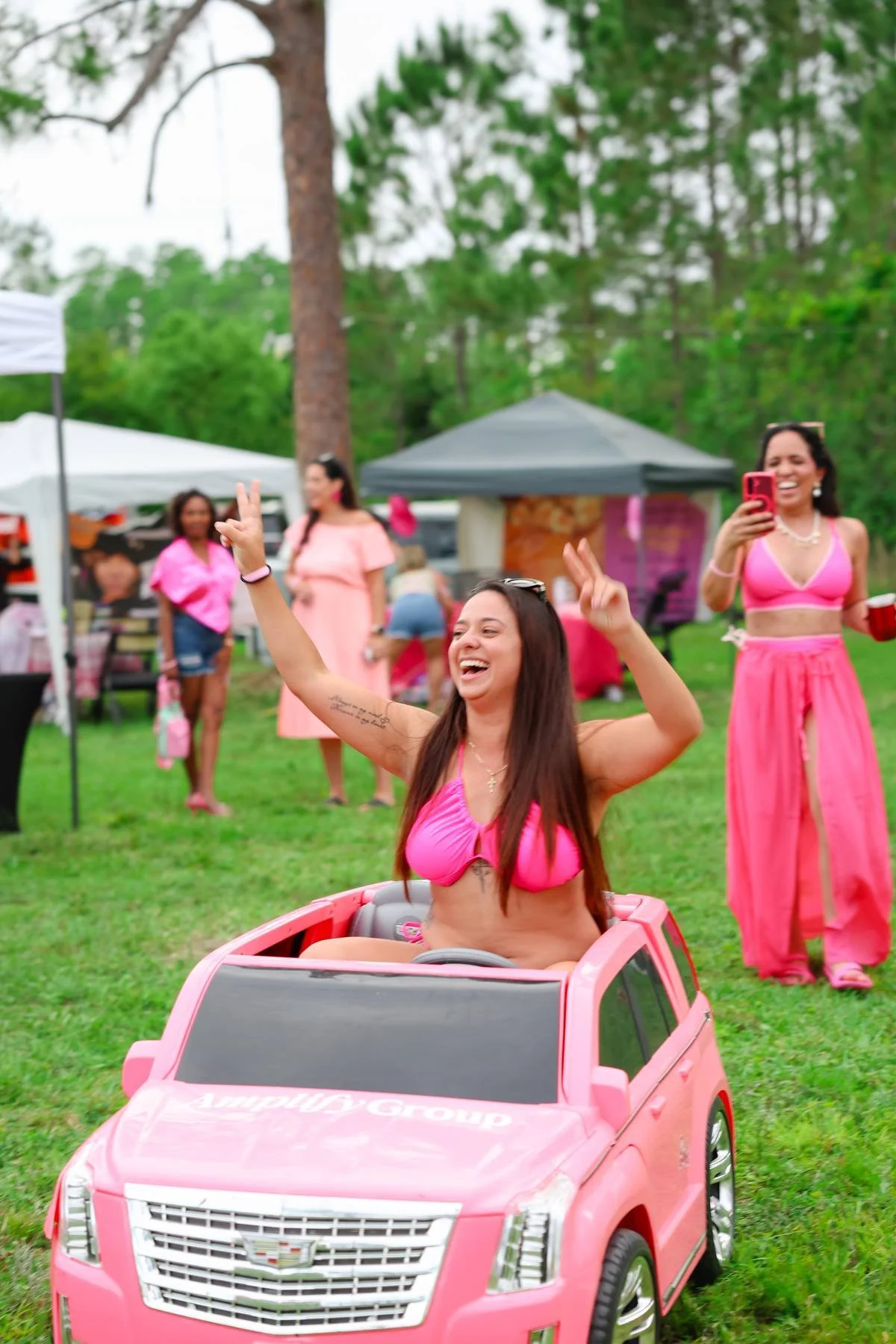 A group of women enjoying outdoor party. One woman in pink bikini is riding a pink toy Cadillac. Others in the background are standing and chatting, some wearing pink outfits. The setting is a grassy area with trees and tents.