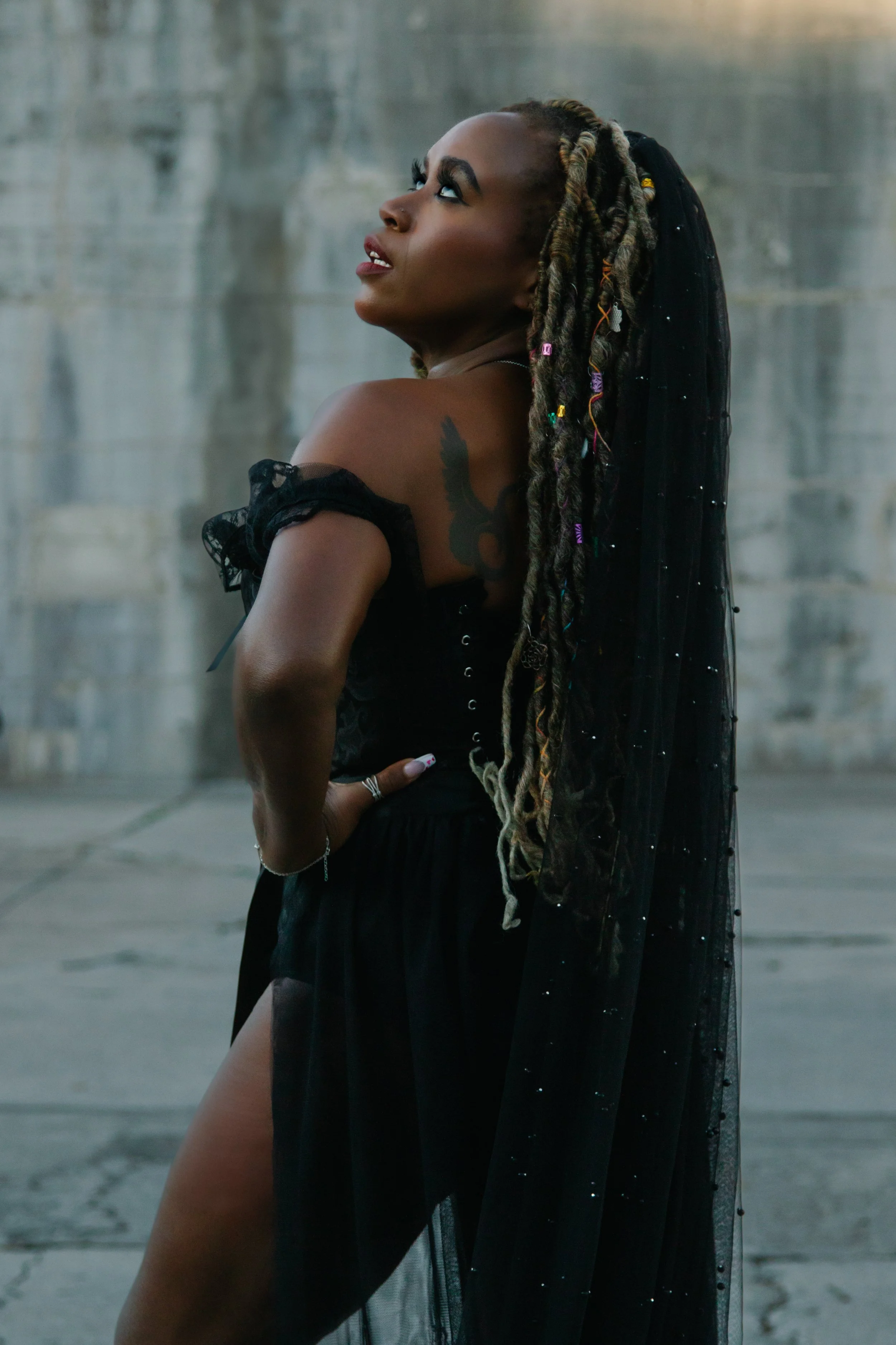 A woman with long dreadlocks and a tattoo on her shoulder, standing outdoors against a concrete wall, wearing a black outfit with a sheer, beaded cape.