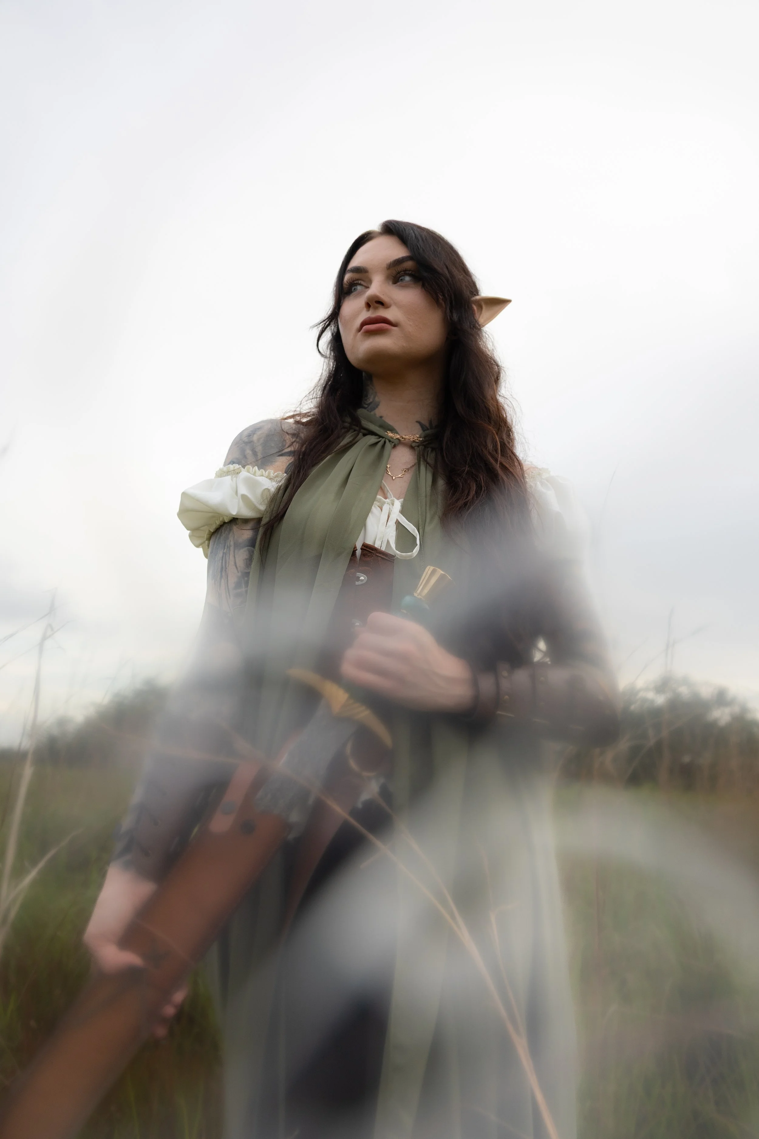 A woman with elf-like ears and tattoos, holding a gun, standing outdoors in a grassy field on an overcast day.