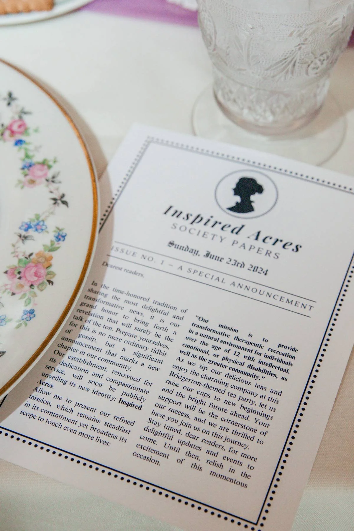 A printed announcement from Inspired Acres Society Papers, dated Sunday, June 23rd, 2024, on a white sheet of paper, with part of a floral patterned plate and a textured glass filled with water visible.
