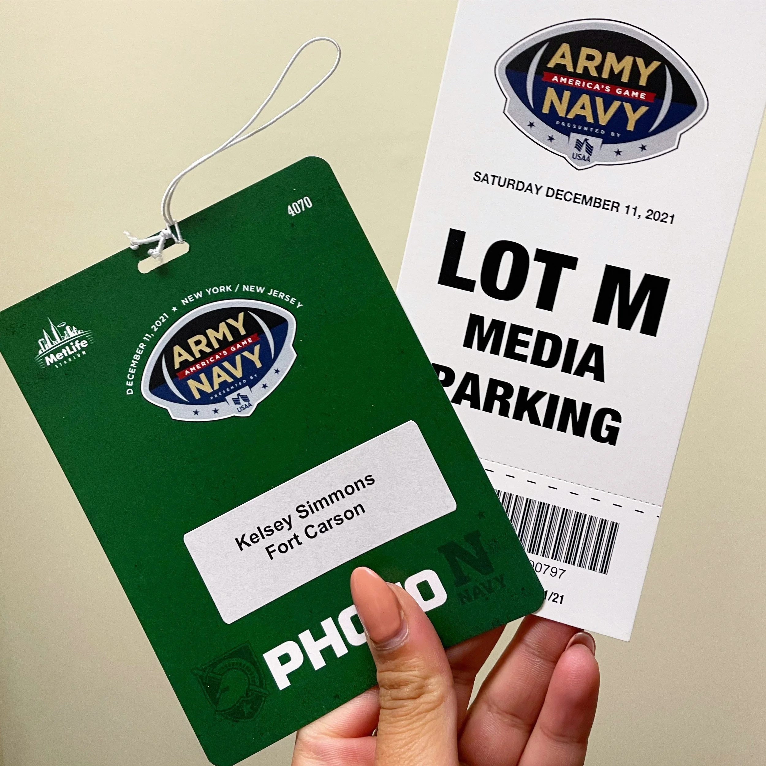 Close-up of a hand holding a green lottery ticket and a white event ticket. The event ticket has the Army-Navy logo with the date December 11, 2021, printed on it.