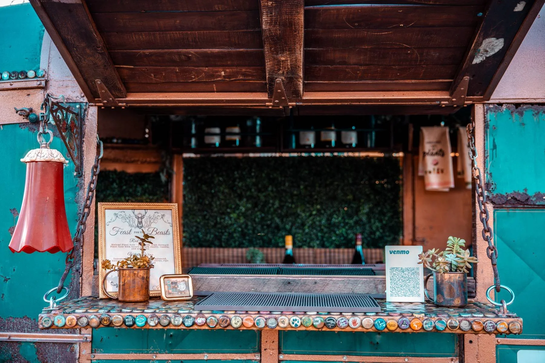 A view of an outdoor bar or food stand with a rustic wooden roof. The bar has a row of bottle caps along the front edge and various decorative items on the counter, including a framed menu, a small plant in a pitcher, and a Venmo QR code sign. Behind