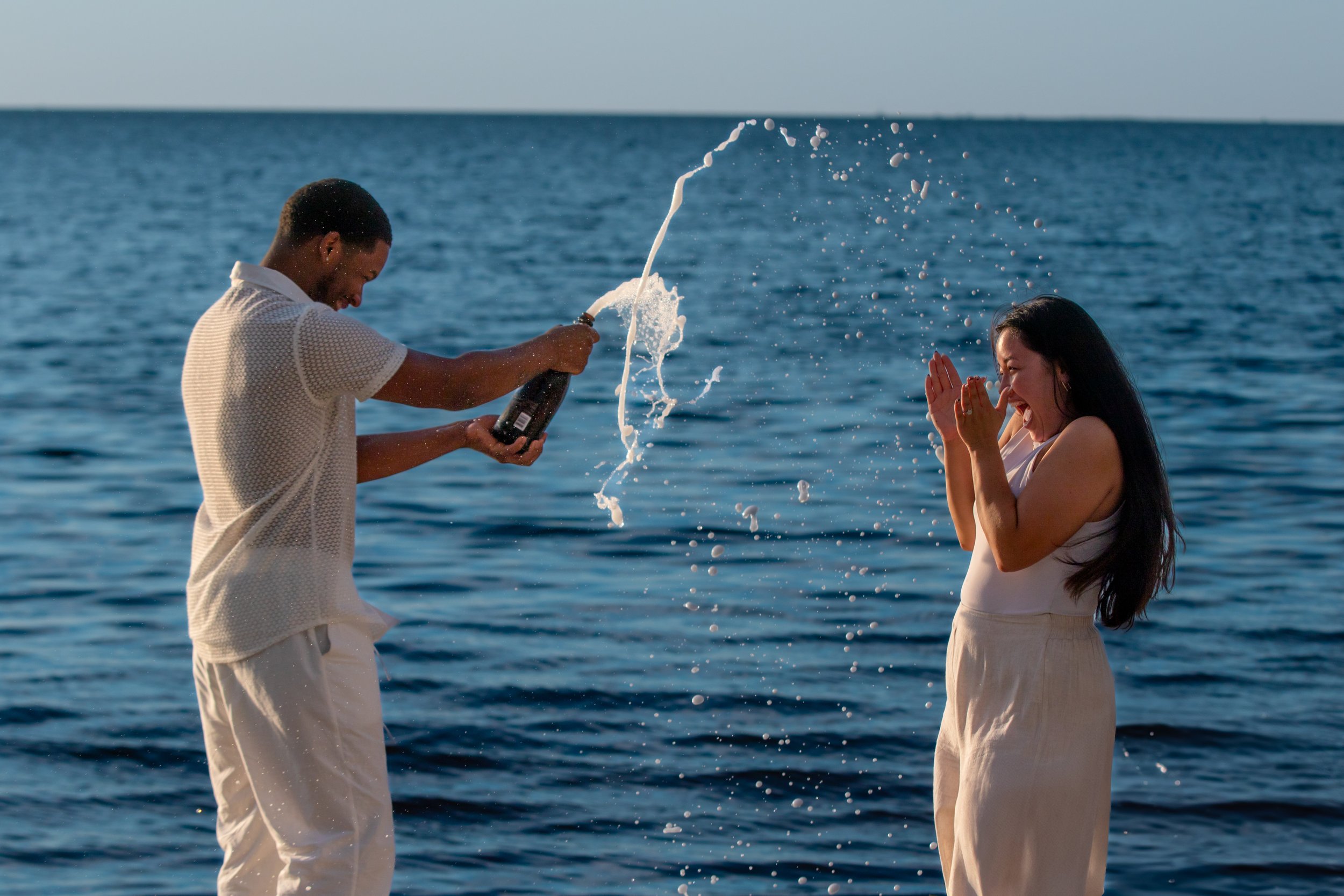 A man and a woman are by the ocean, laughing and playing with champagne, with the man spraying champagne into the woman's face.