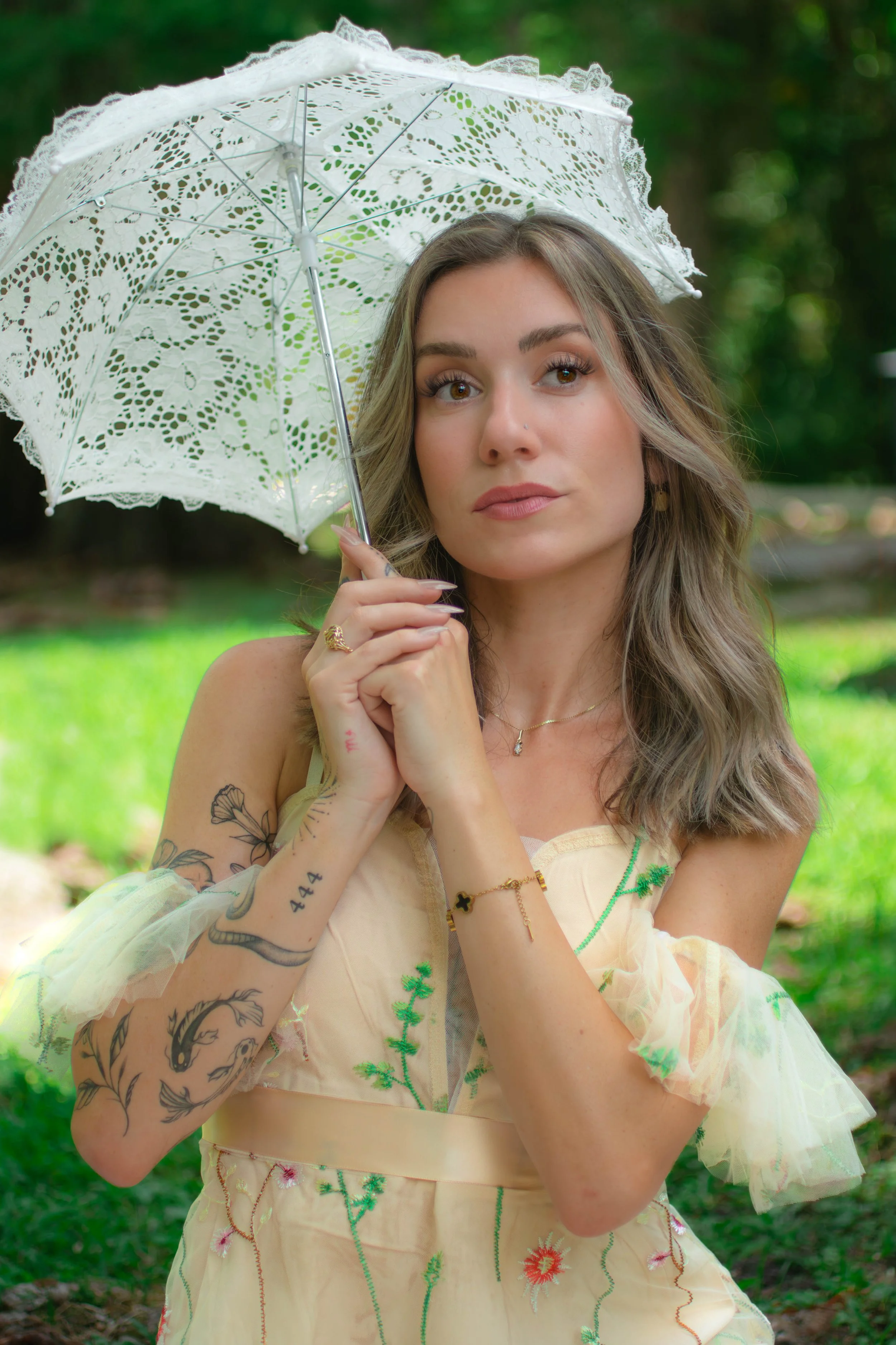 A woman with long wavy blonde hair holding a white lace parasol outdoors with greenery in the background. She has tattoos on her left arm, light makeup, and is wearing a light-colored dress with green and pink embroidery.
