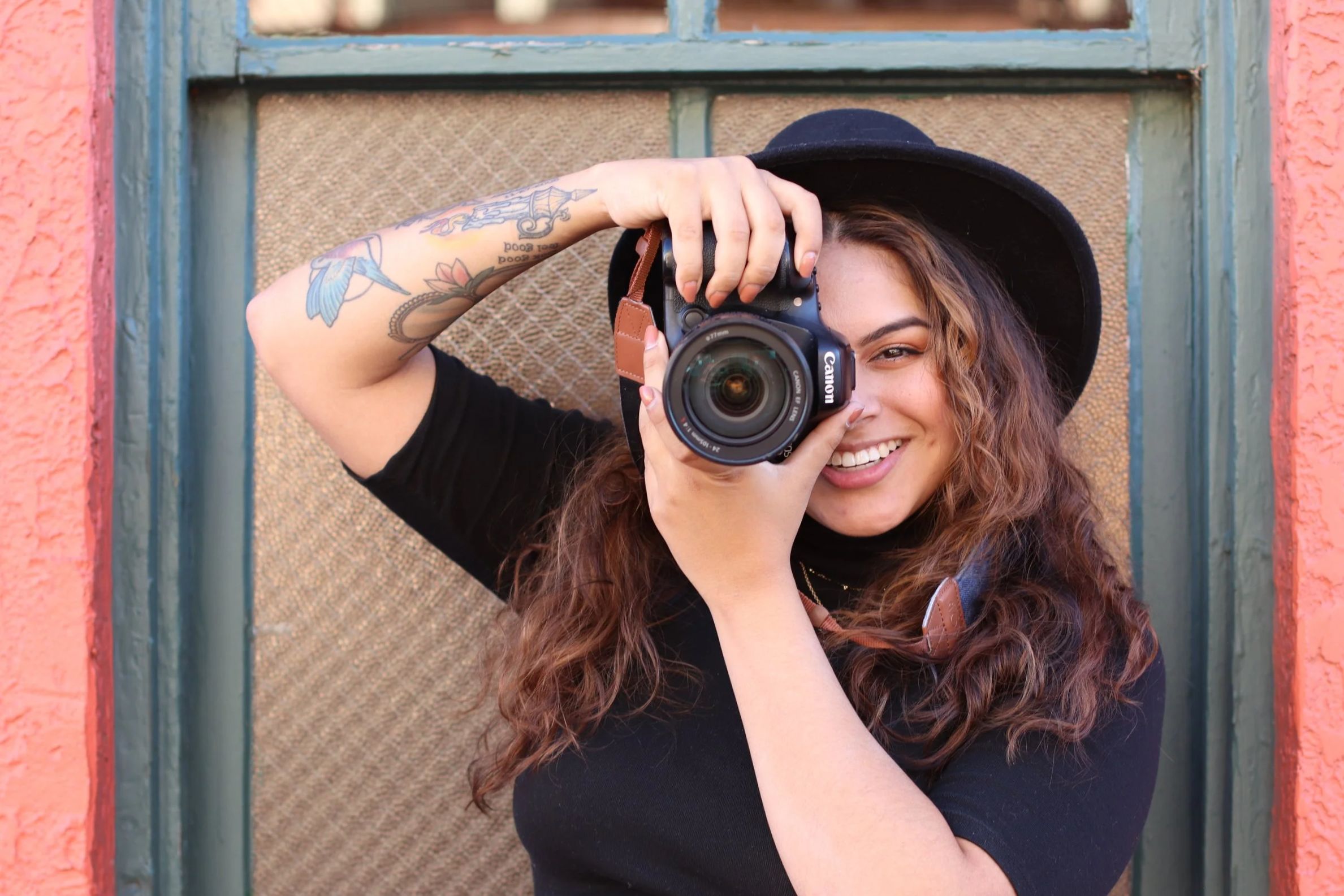 A woman with curly hair and tattoos on her arm is holding a camera and smiling. She is wearing a black wide-brimmed hat and a black shirt, and standing in front of a colorful wall with blue window trim.
