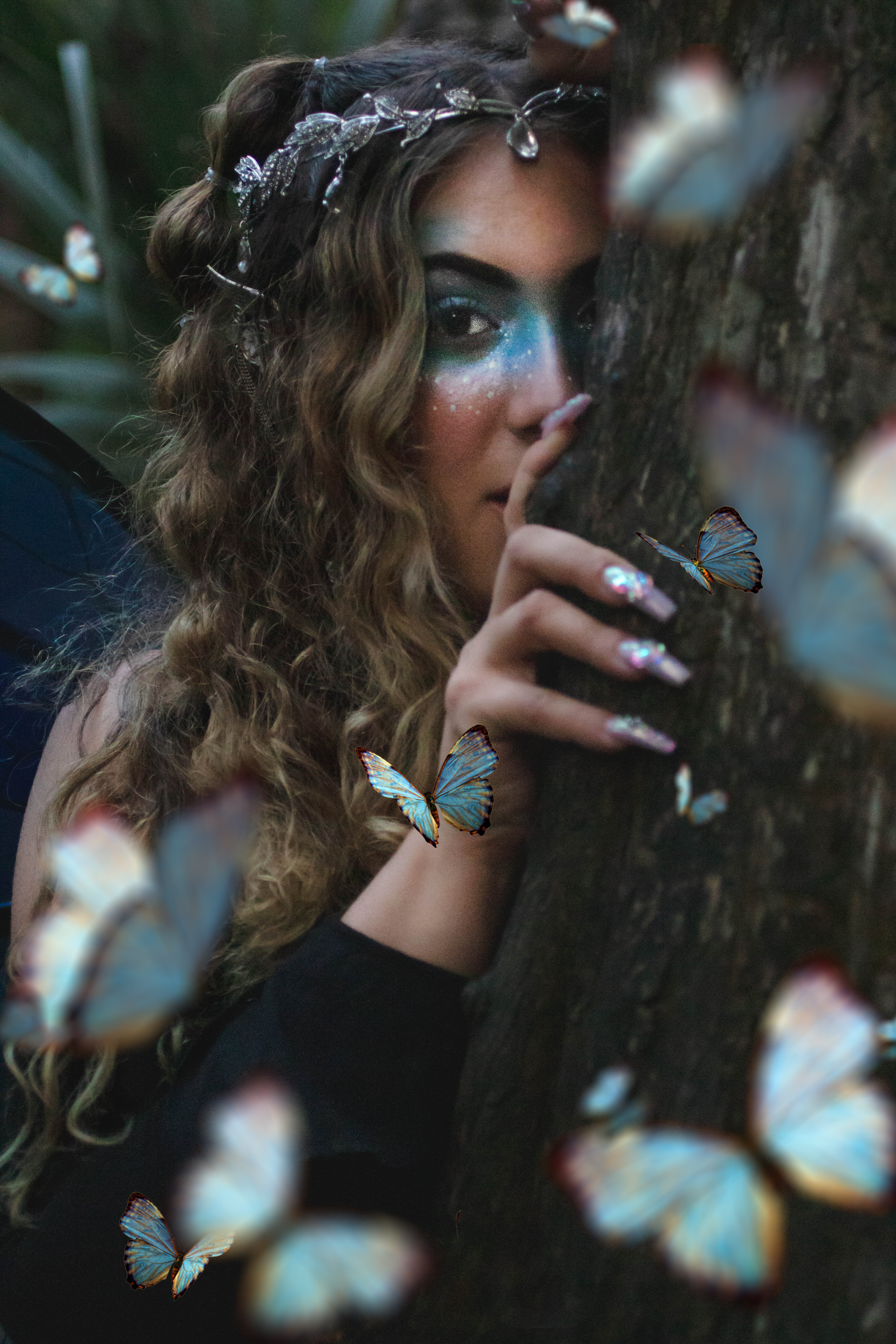 A woman with wavy hair and a silver butterfly headband poses behind a rough tree trunk, touching it with her hand. Her face is decorated with galaxy-inspired makeup, and she is surrounded by blue butterflies flying around her in a mystical environmen