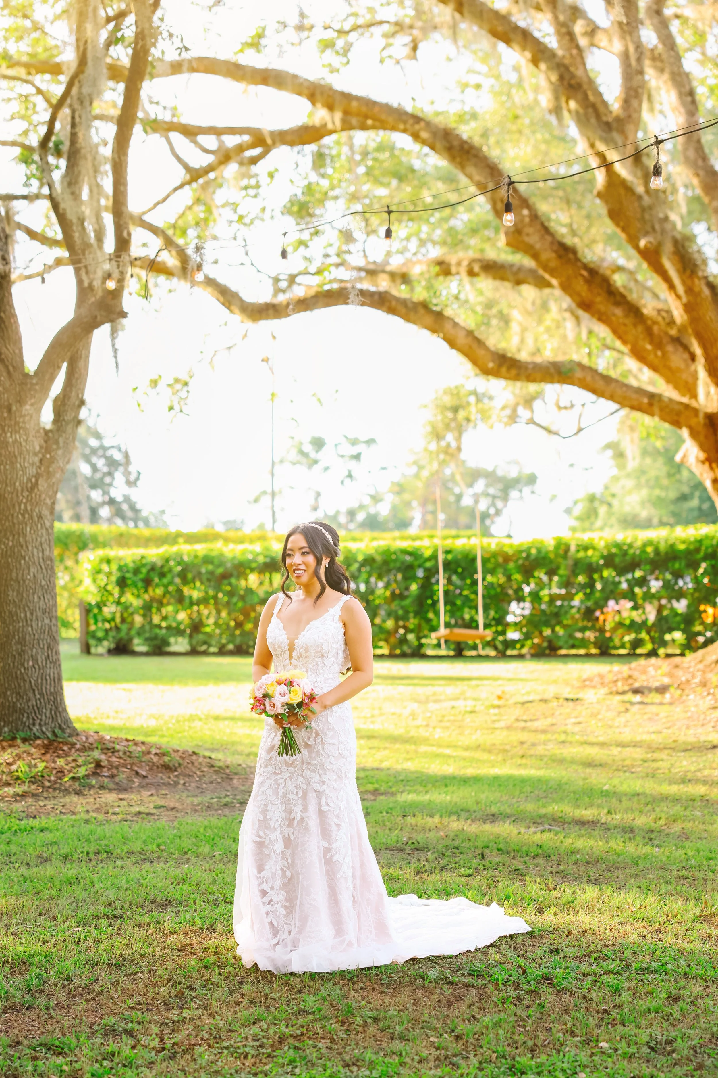 Bride in white wedding gown holding a bouquet of flowers standing outdoors on grass with large trees and string lights in the background.