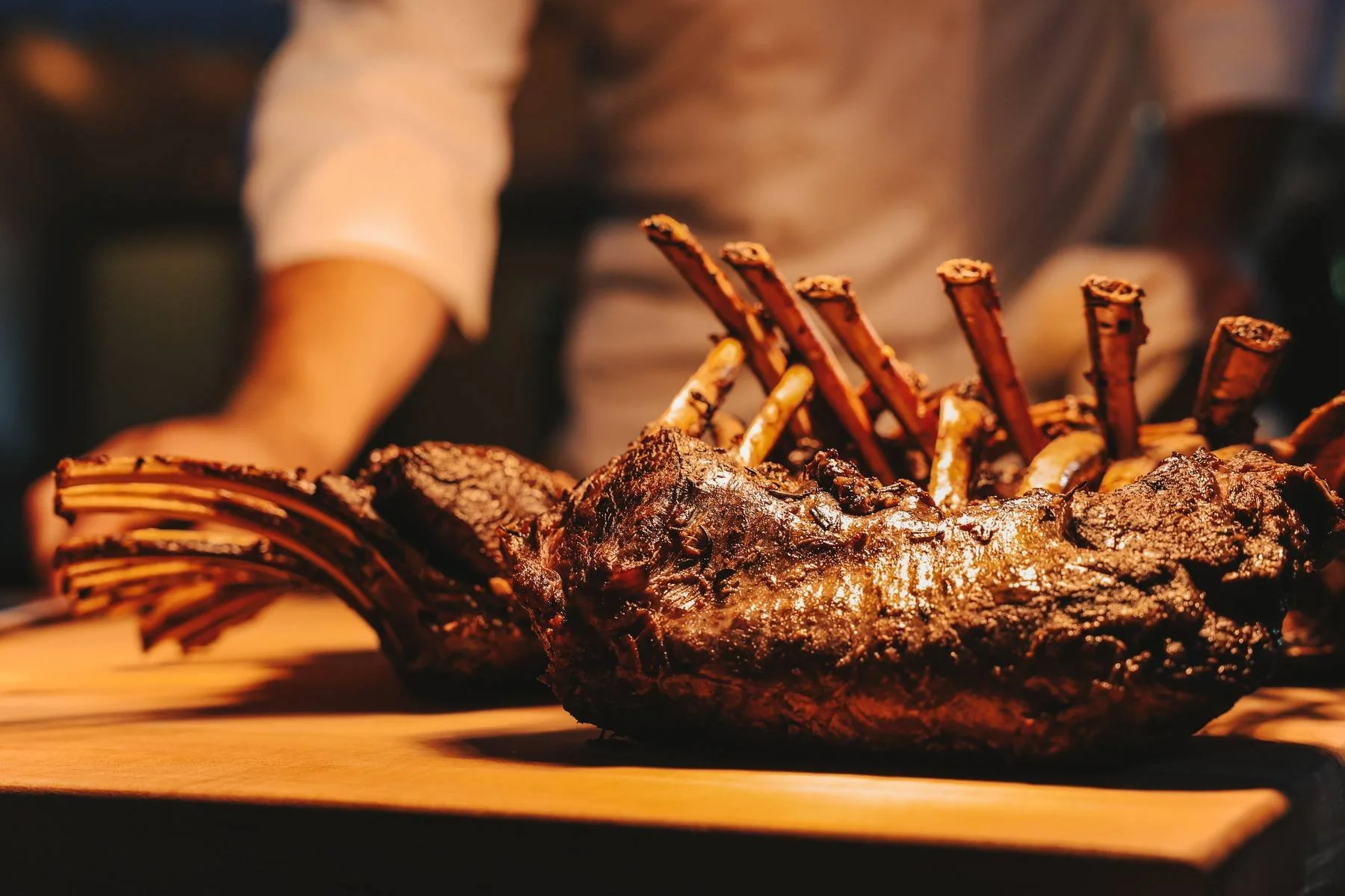 Close-up of a roasted rack of lamb with crispy breaded edges on a wooden surface, with a blurred person in the background.