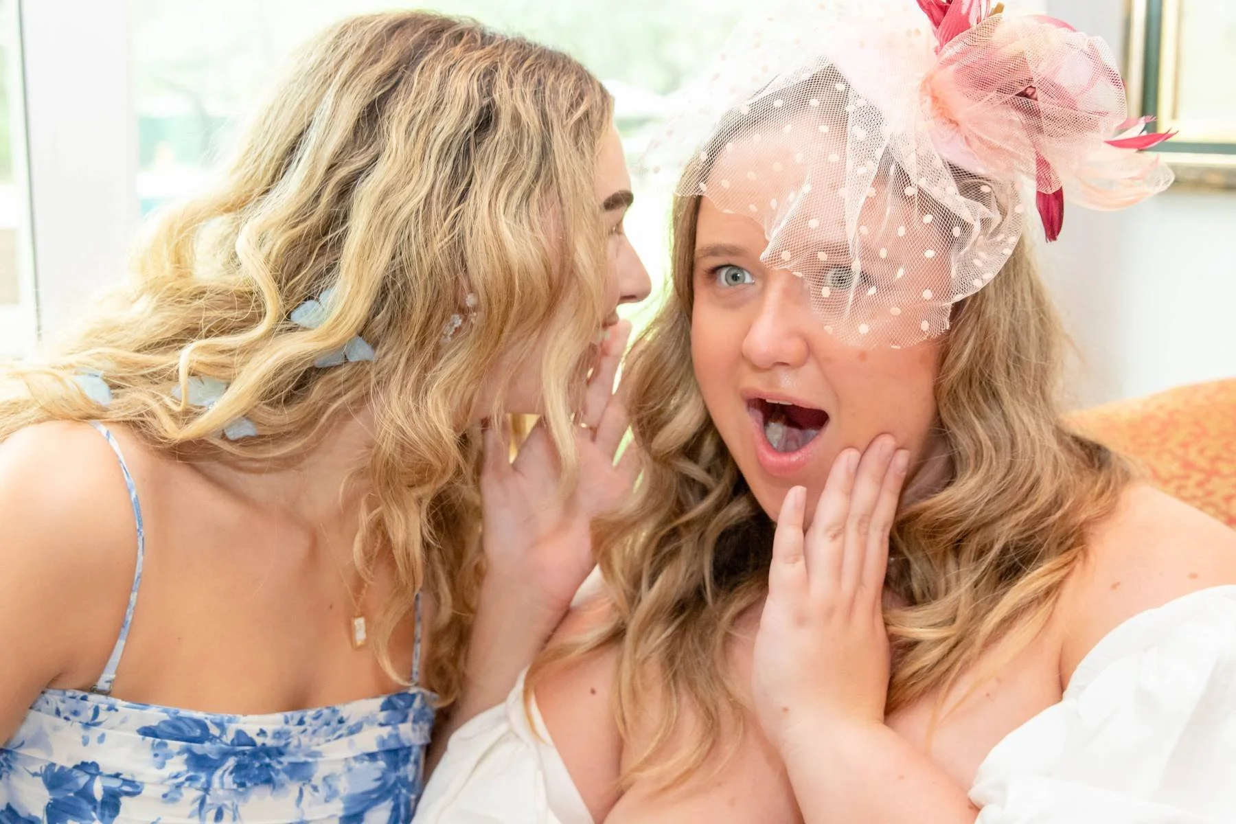 Two women with blonde curly hair having a secret conversation. One woman is whispering while the other looks surprised and holding her cheek, wearing a pink fascinator with tulle.