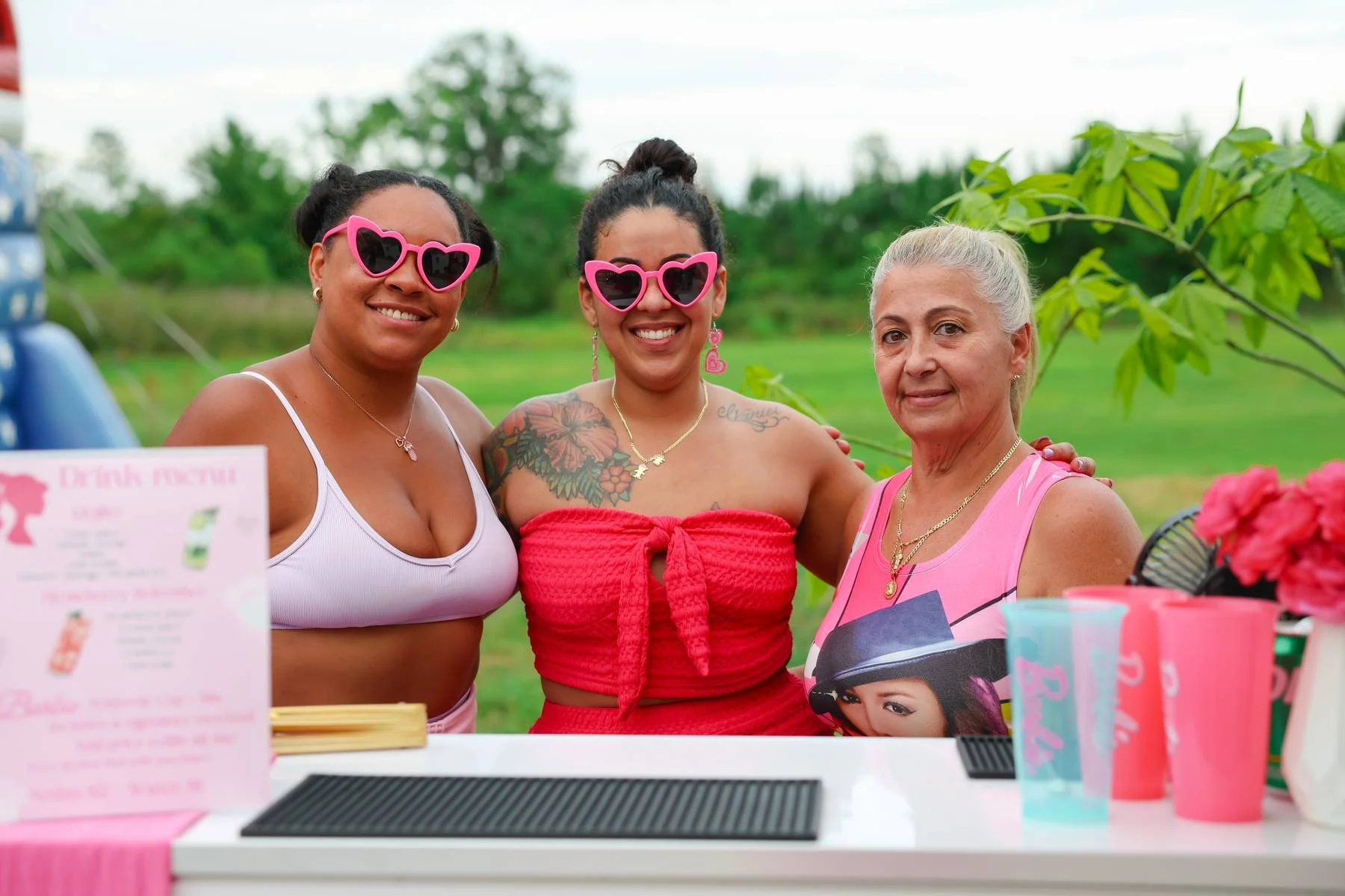 Three women standing outdoors at a market or fair, smiling and wearing sunglasses with heart-shaped frames. They are dressed in casual summer clothing, with two in pink tops and one in a red top. There are green trees in the background, and items suc