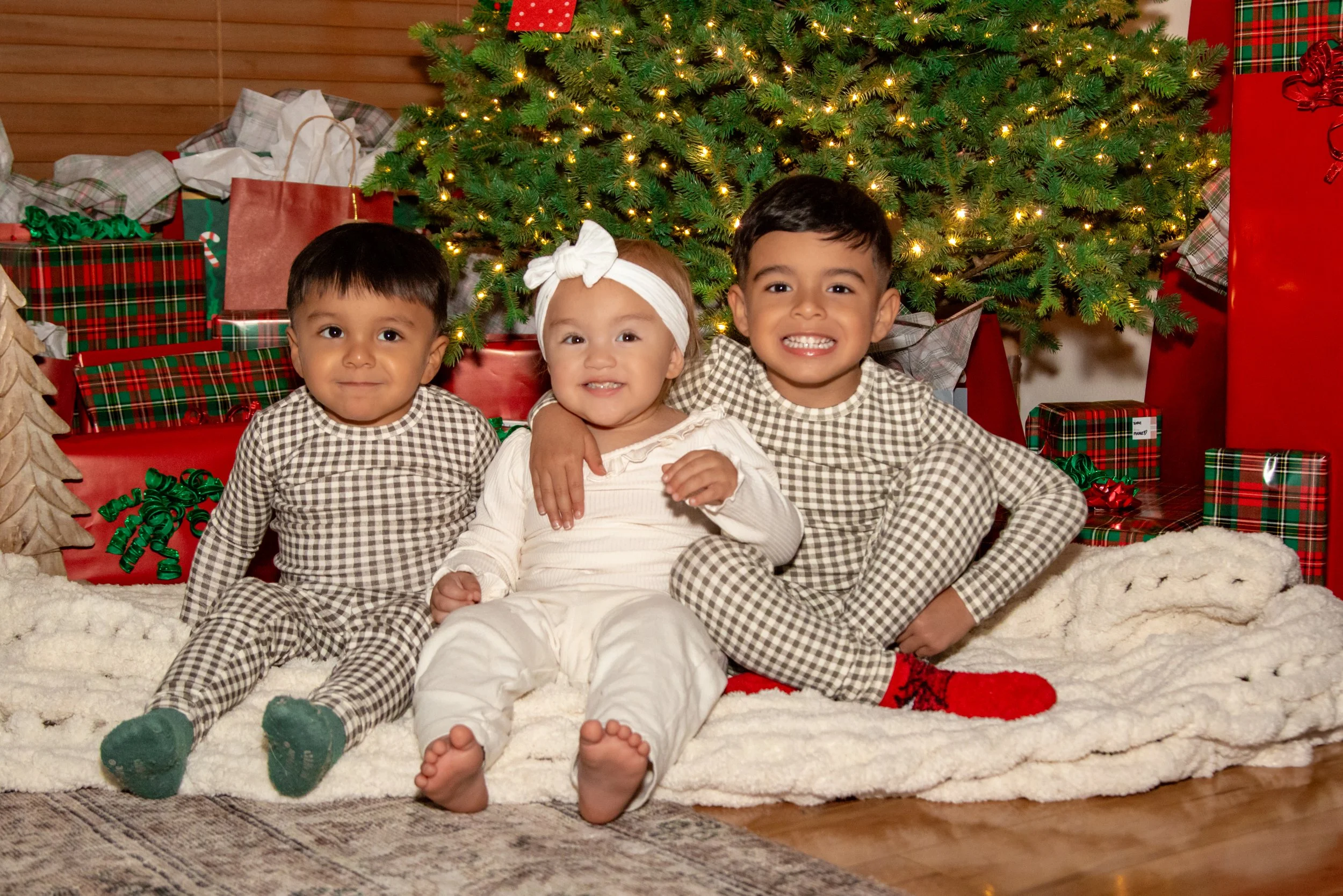 Three young children sitting on a fuzzy white rug in front of a decorated Christmas tree with wrapped presents behind them.