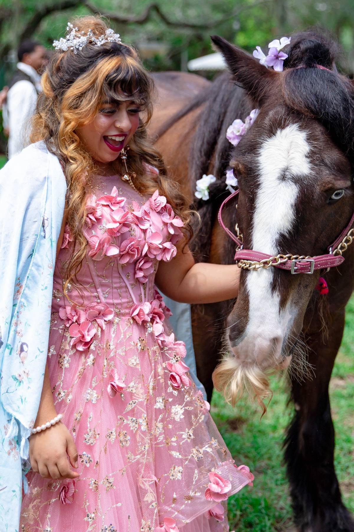 A woman in a pink dress adorned with pink flowers and gold embroidery, smiling excitedly, stands next to a dark brown horse decorated with purple and white flowers in an outdoor setting with trees. She is wearing jewelry and a crown, with a light blu