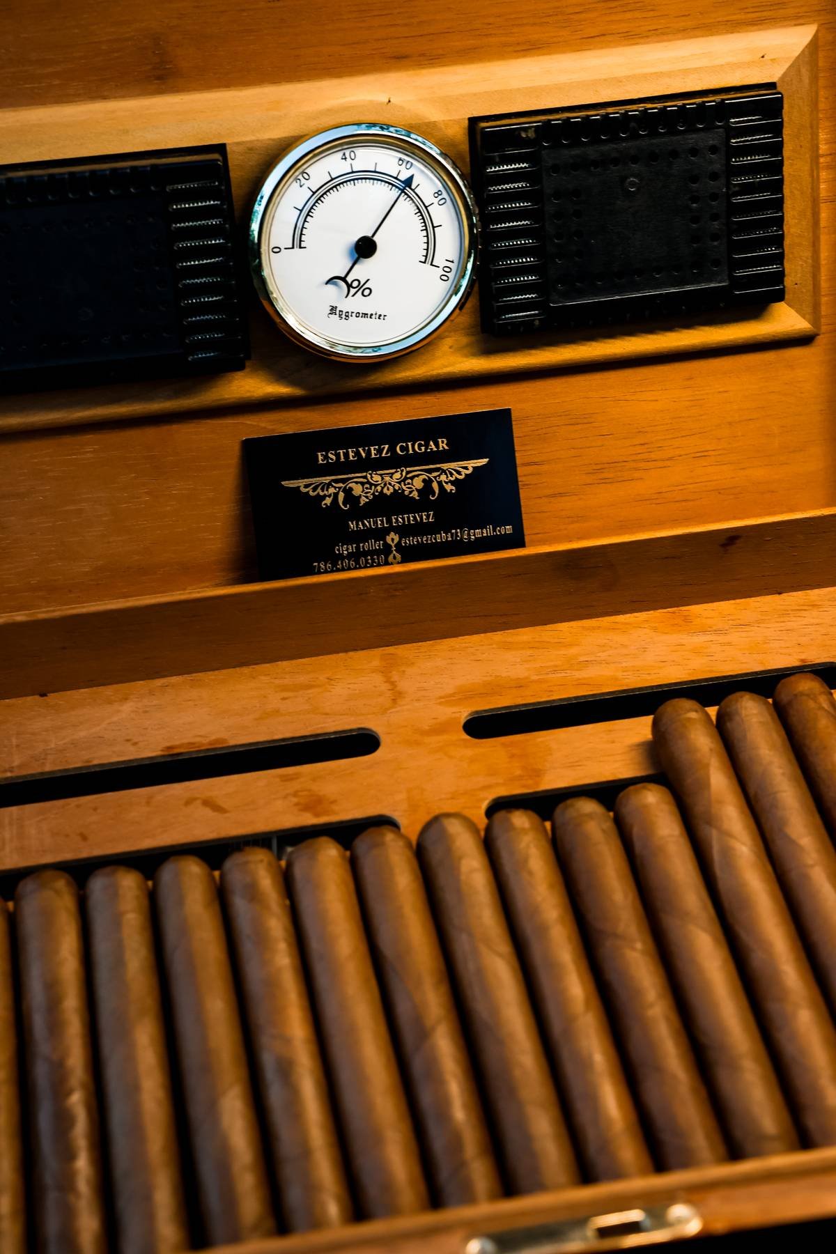 Close-up of a wooden cigar humidor with a hygrometer and humidor drawers filled with cigars. Business card with contact information for Estevez Cigar and Manuel Estevez.