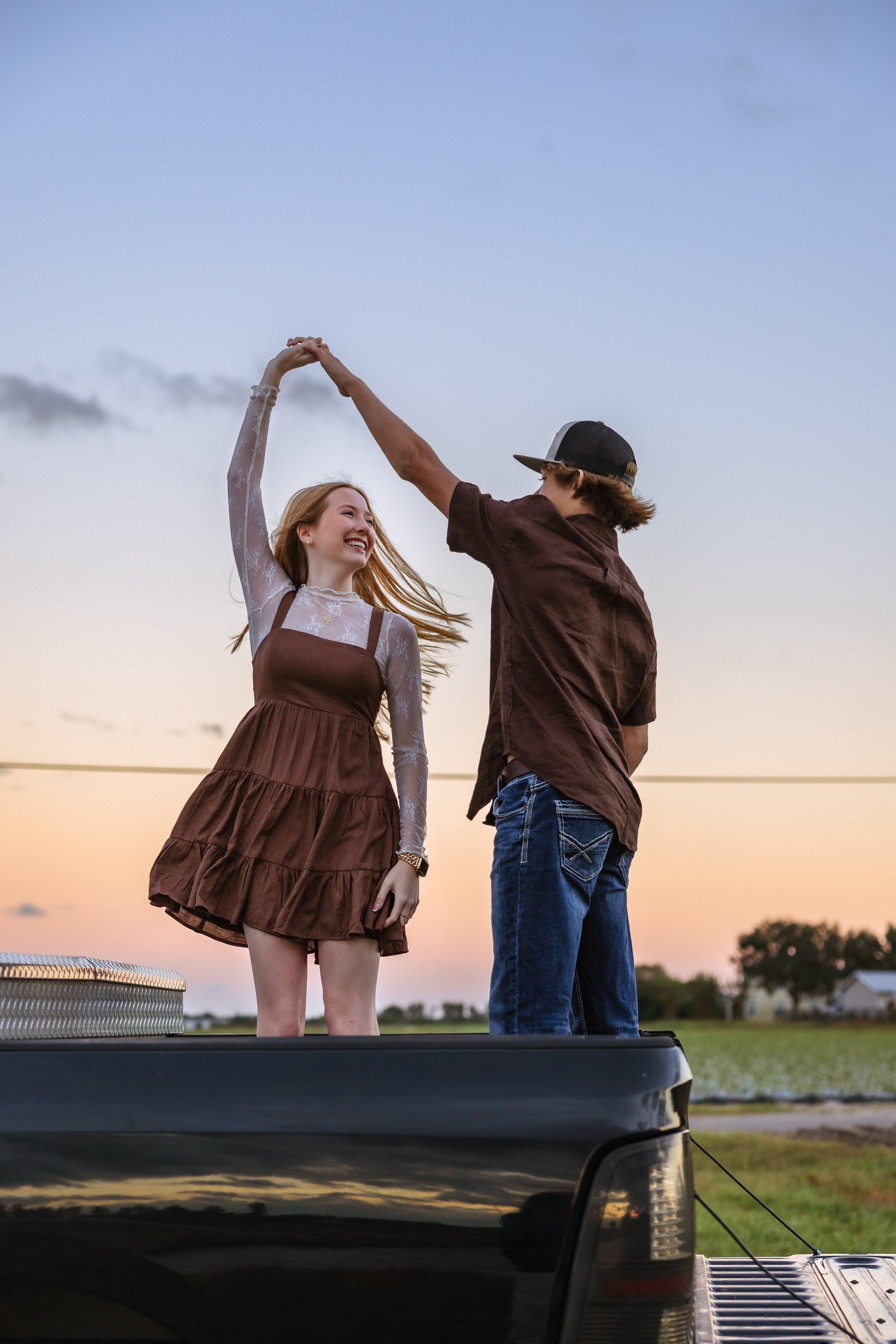 A young woman and a young man dancing on the back of a pickup truck during sunset. The woman is smiling and wearing a brown dress with lace sleeves, while the man is wearing a brown shirt, jeans, and a black cap. They are holding hands and facing eac