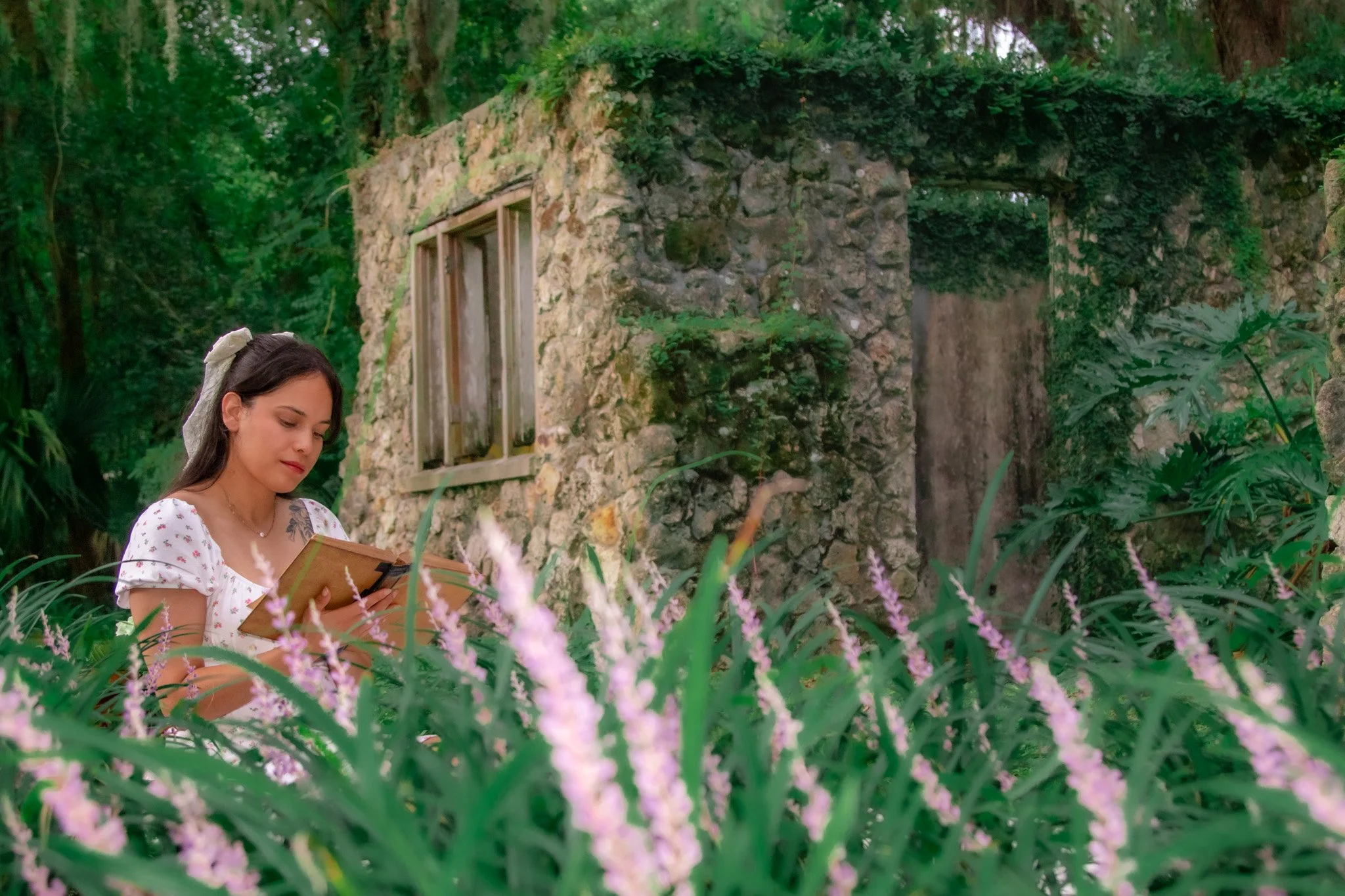 A young woman with dark hair and a white hair ribbon reads a book or notebook in a lush garden surrounded by pink flowers, green foliage, and an old stone building covered with vines.