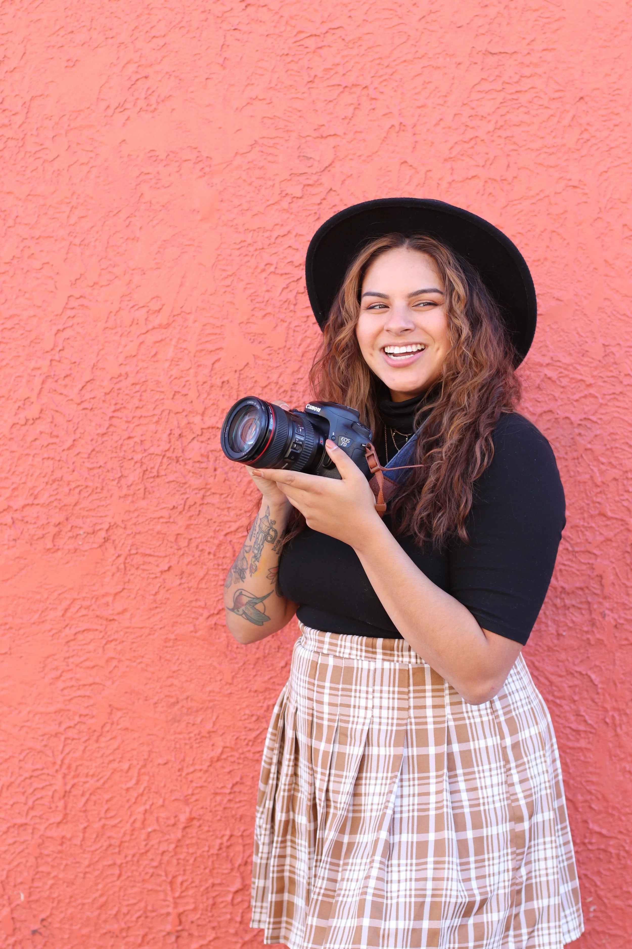 A young woman with curly brown hair, wearing a black wide-brimmed hat and a black top, holding a camera and smiling in front of an orange textured wall.