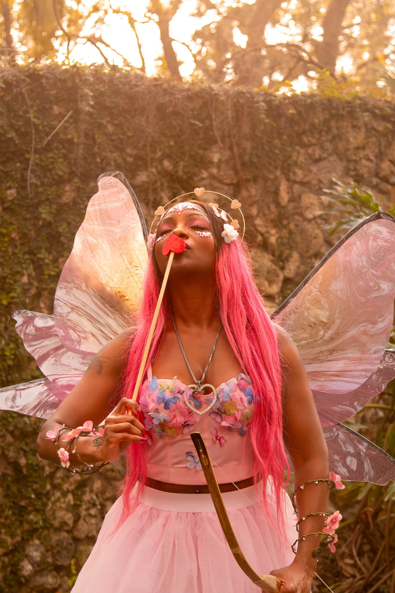 A woman dressed as a fairy or butterfly with pink hair, fairy wings, and floral accessories, holding a heart-shaped wand and standing outdoors with trees and sunlight in the background.