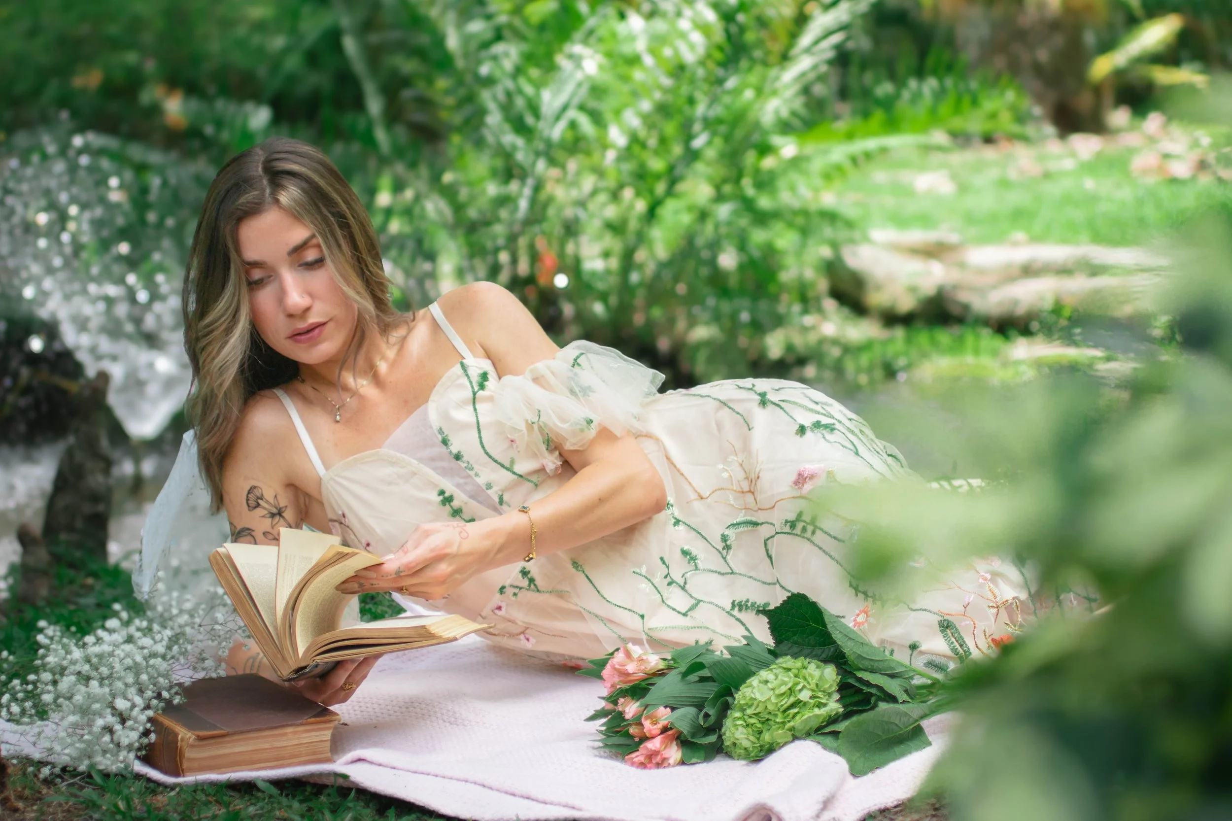 Woman in a white dress with floral embroidery reading a book while lying on a blanket outdoors surrounded by greenery and flowers.