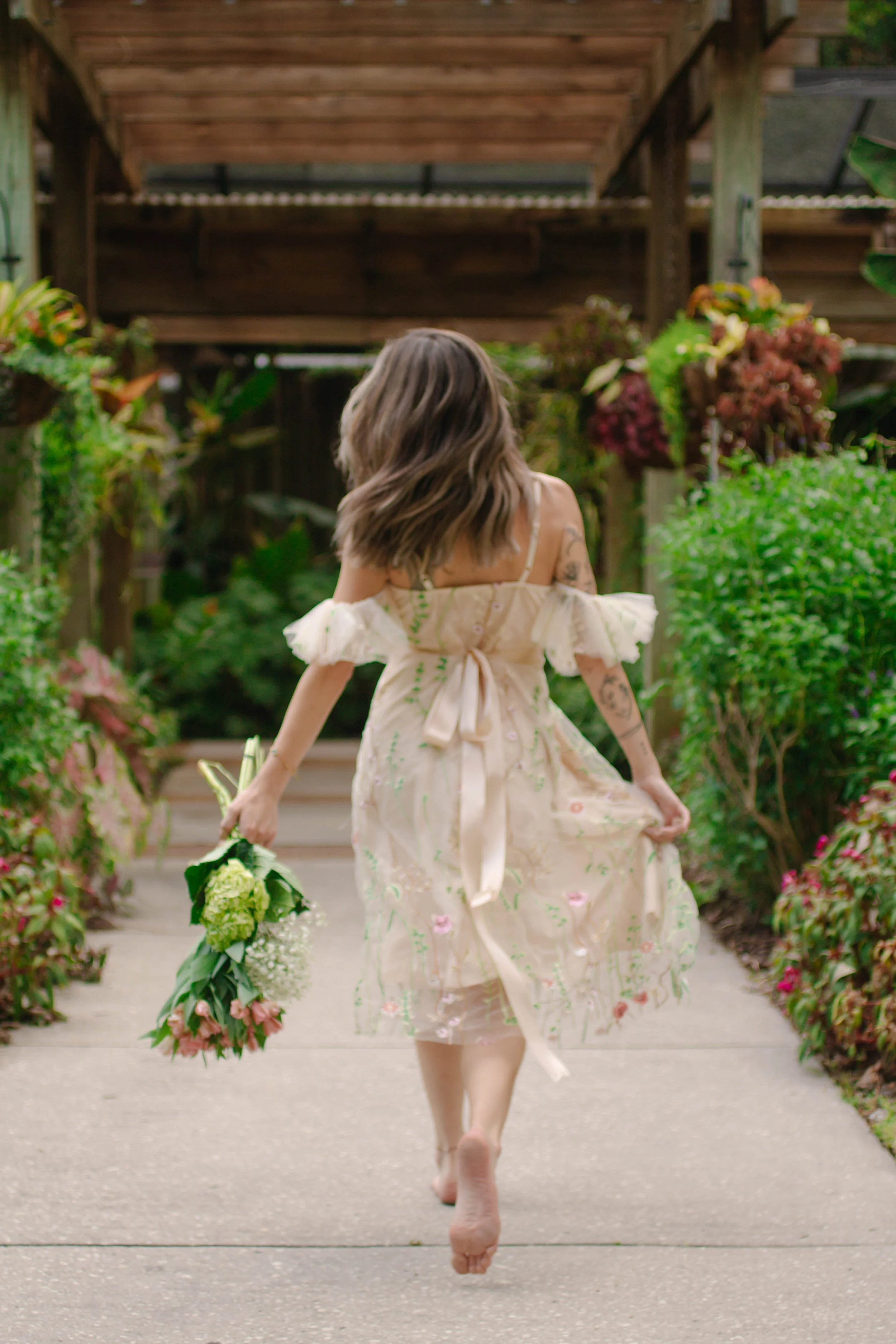 A woman with shoulder-length hair walking barefoot on a sidewalk surrounded by lush greenery and flowers, holding a bouquet of white and pink flowers.