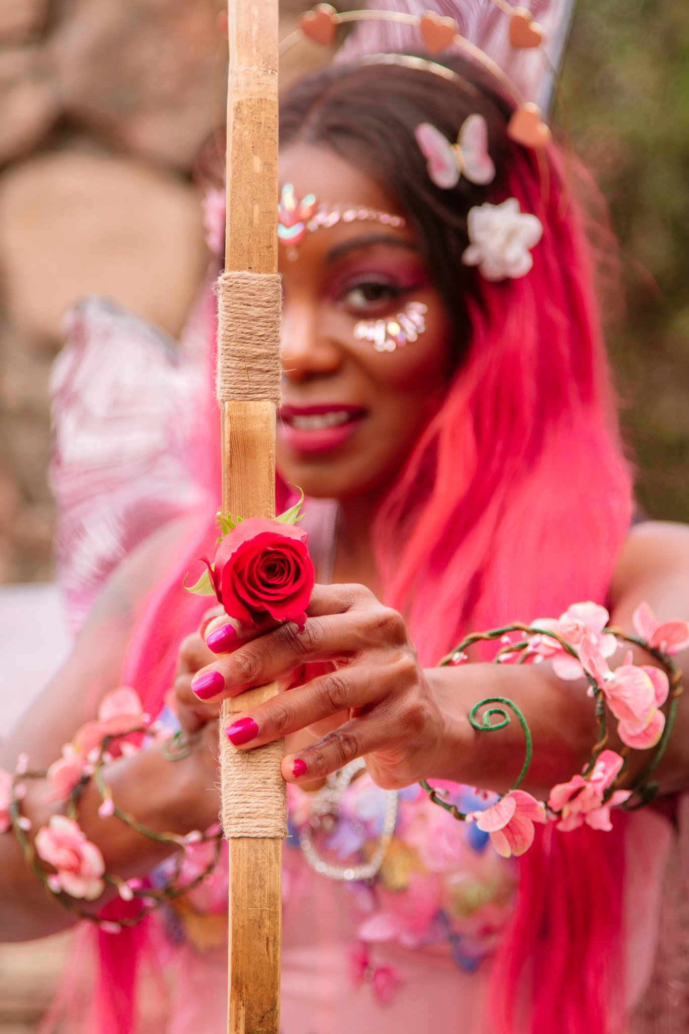 A woman with pink hair and floral makeup is holding a staff wrapped in twine, wearing a flower and butterfly headband, and adorned with flowers on her arms and shoulders, outdoors with a blurred background.