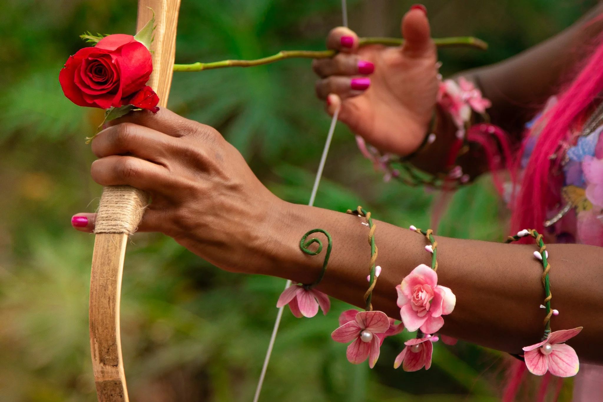 A person holding a bow and arrow, with a red rose attached to the arrow shaft. The person has pink painted nails, bracelets, and wears pink and purple clothing, with a blurred green background.