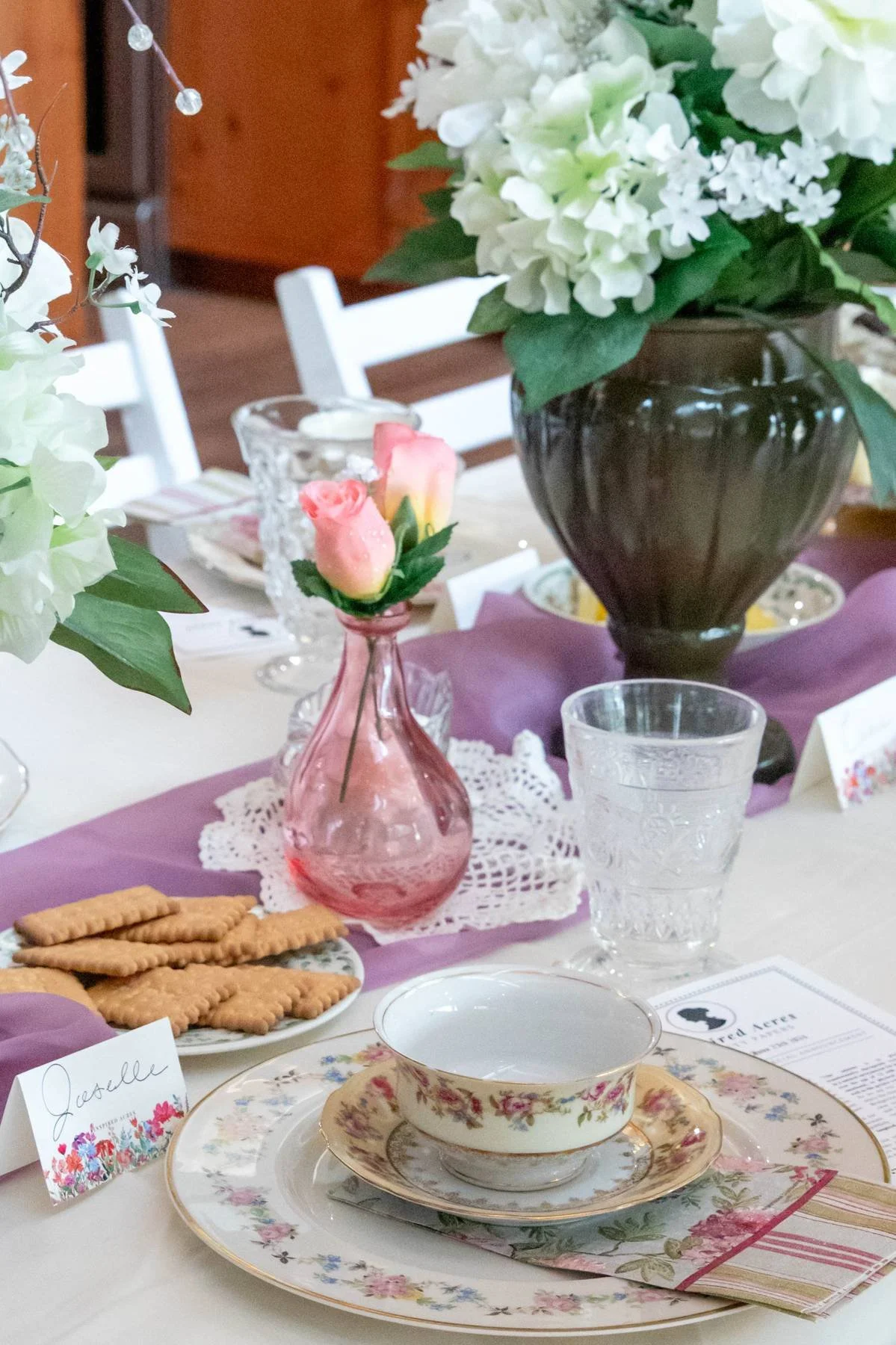 A table set for an elegant meal with floral arrangements, a pink vase with roses, a plate of crackers, fine china teacups, and glasses, with a purple table runner and white chairs in the background.