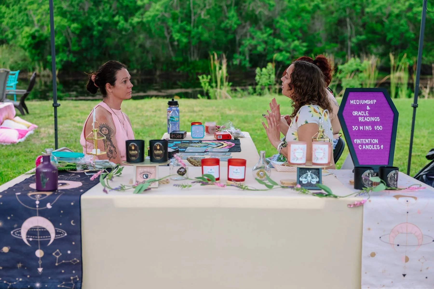 Two women sit across from each other at a table outdoors by a river, engaged in a tarot reading. The table is decorated with celestial-themed items, candles, and tarot cards. A purple sign advertises mediumship, oracle readings, and candles.