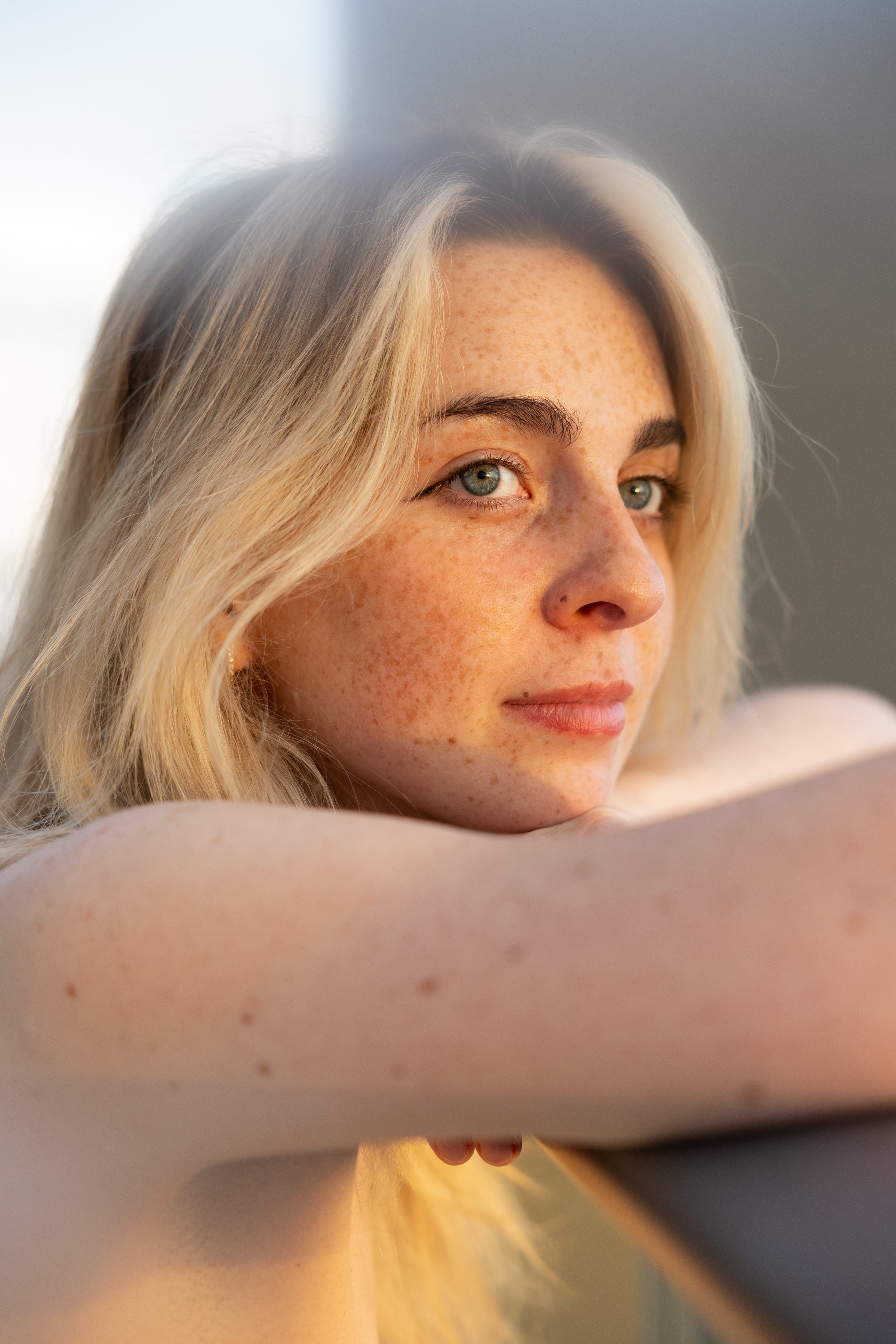 A young blonde woman with freckles resting her arms on a surface and looking into the camera with blue eyes.