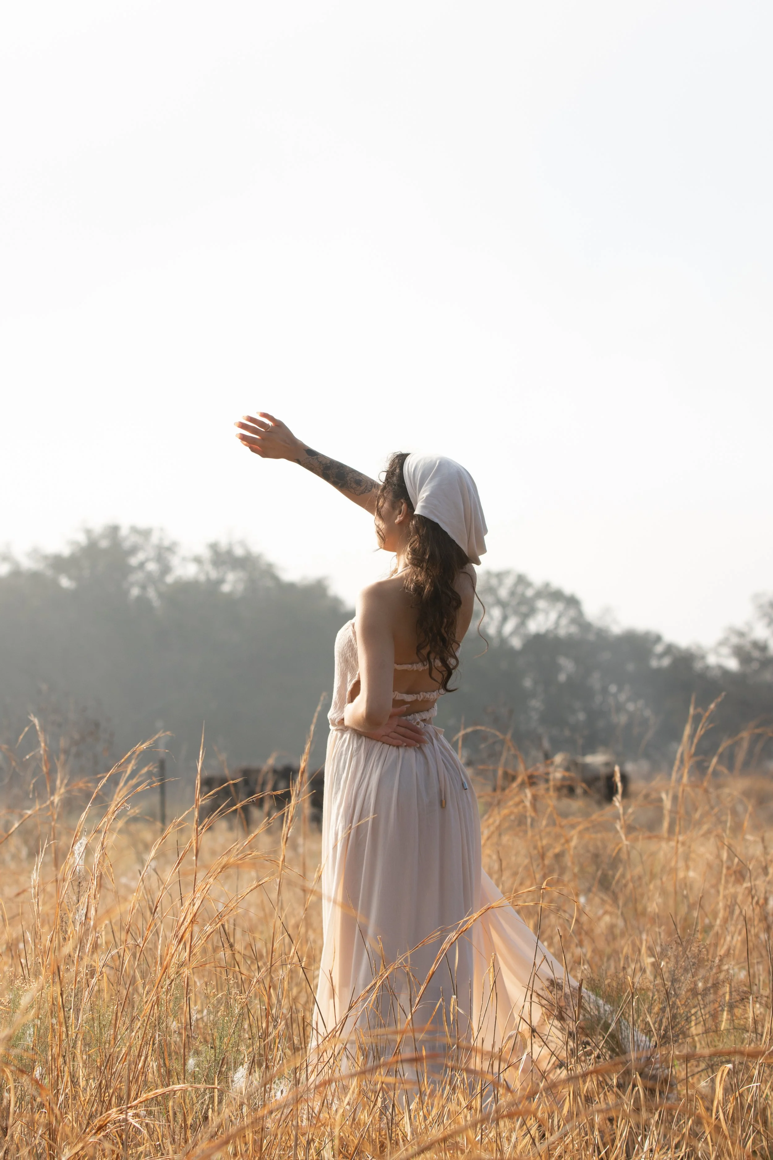 A woman in a flowing beige dress with long curly hair and a white head covering standing in a field of tall dried grass, with trees in the background under a bright sky.