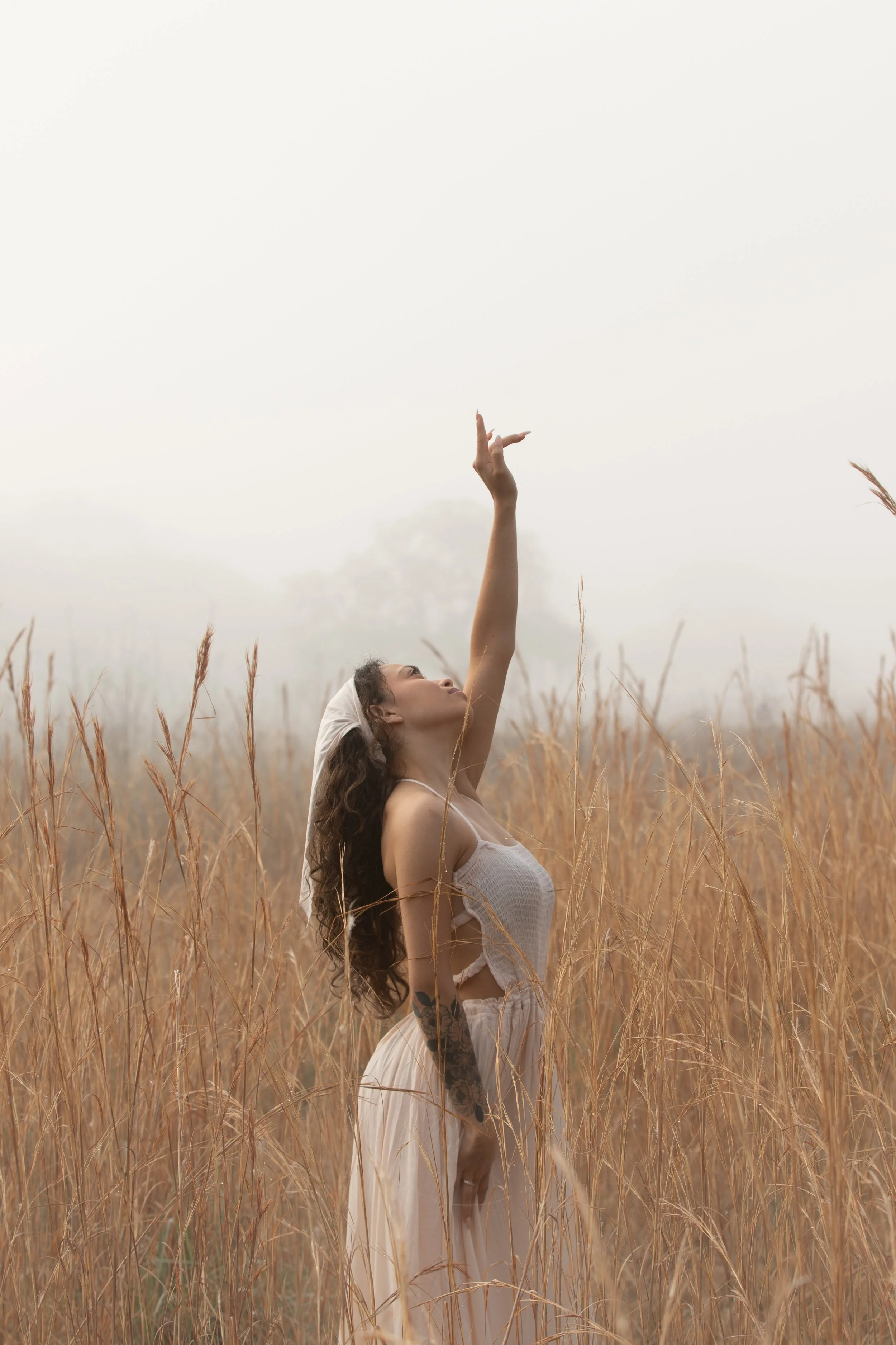 A woman in a white dress and headscarf standing in a field of tall, dry grass, with her arm raised upward. A cloudy sky is in the background.