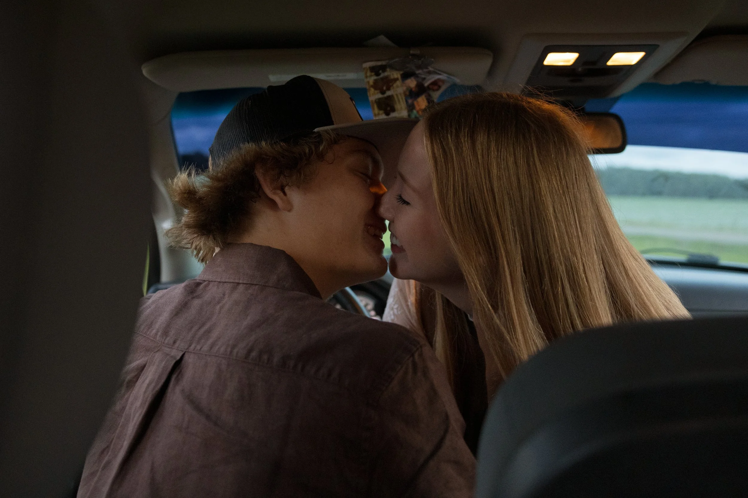 A young man and a young woman are sharing a kiss inside a car, with the horizon and sky visible through the windshield.