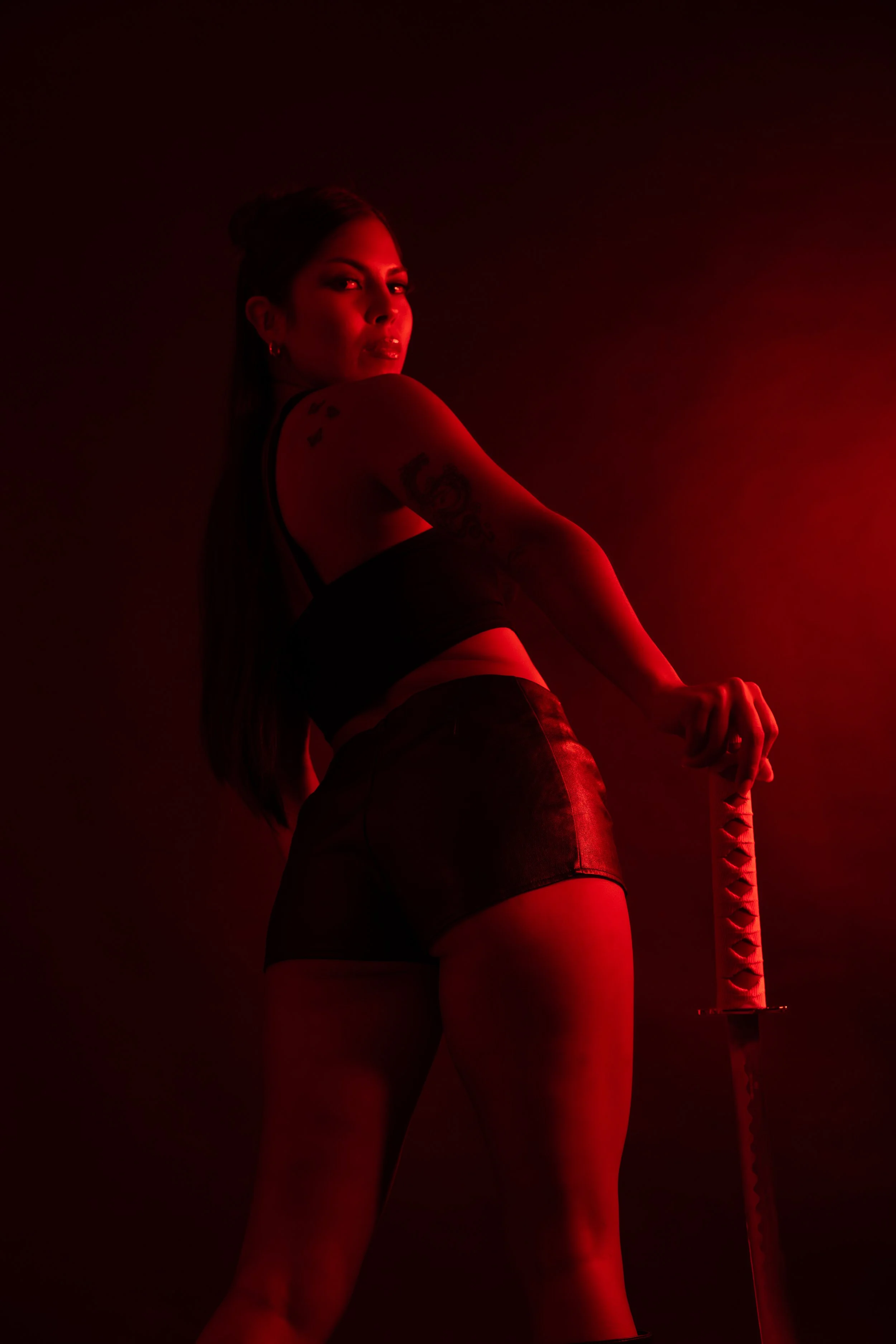 A woman with tattoos on her arm is standing in a dark room illuminated by red light, holding a katana sword.